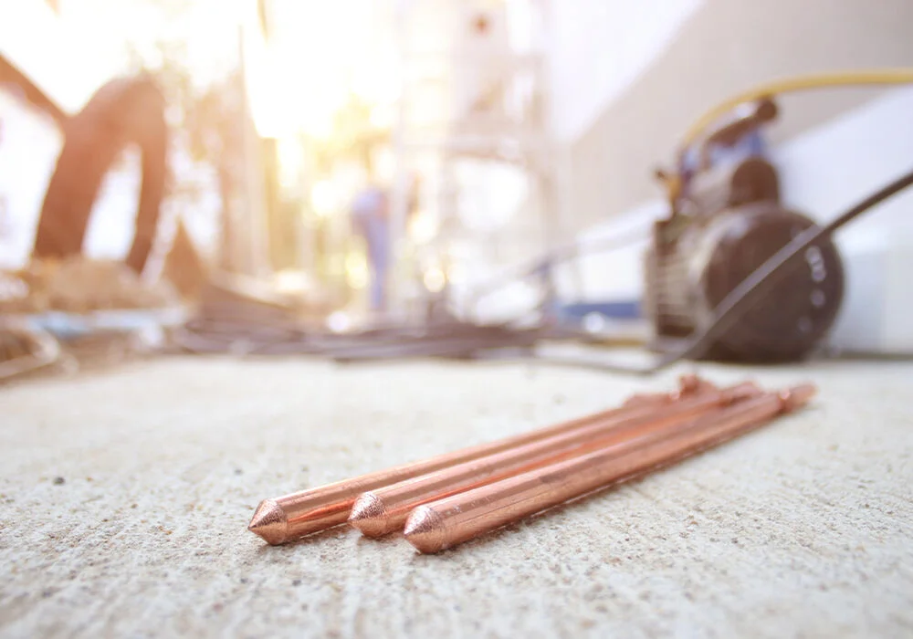 Three copper-coated grounding rods lie on a concrete surface in an outdoor construction area.