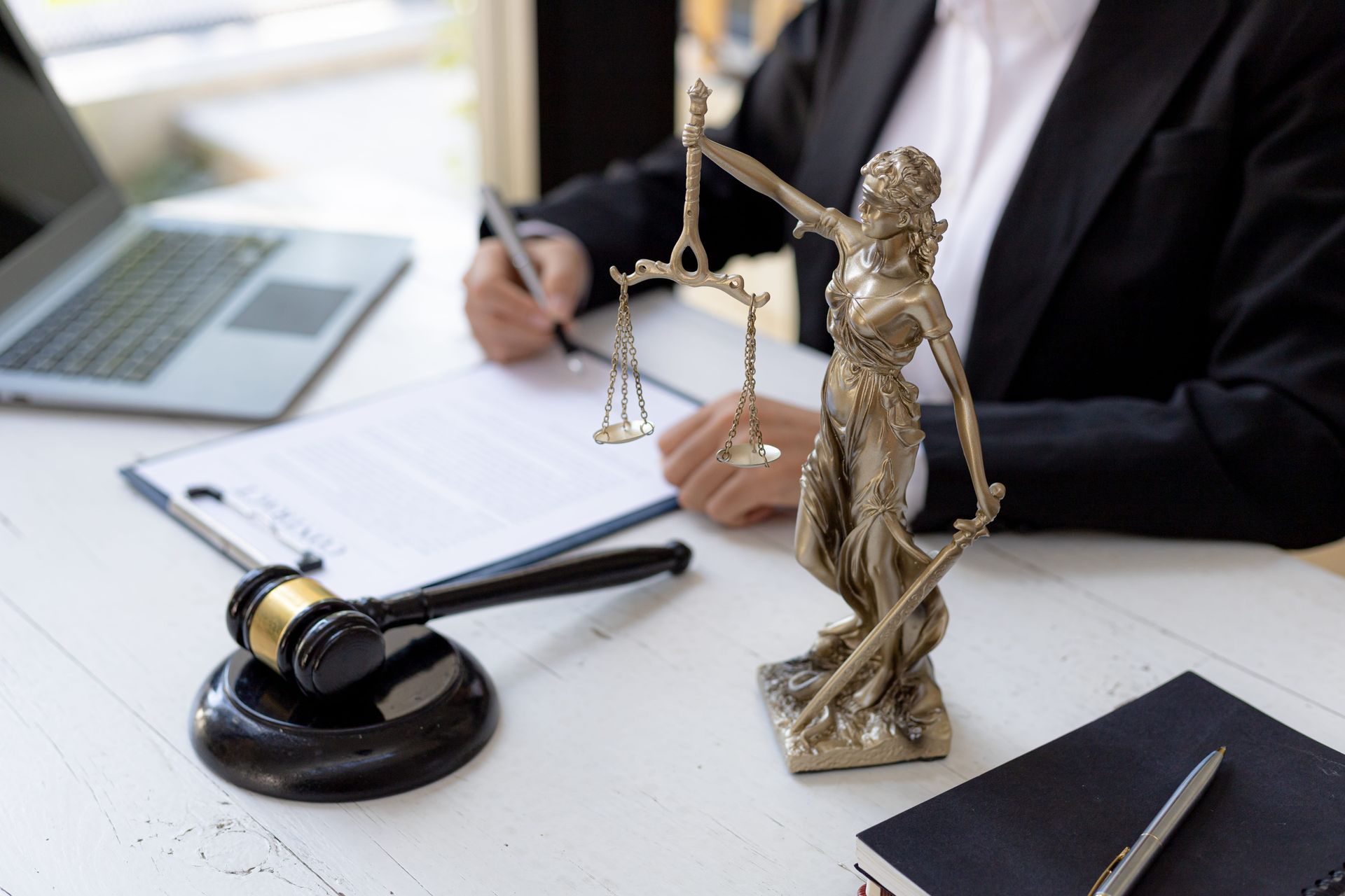 Law office desk with gavel, Lady Justice statue, scales, and a lawyer working on a laptop