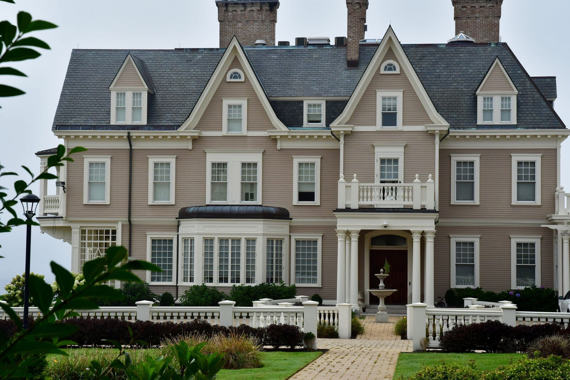 Large beige mansion with gray roof and manicured front garden, seen from the driveway