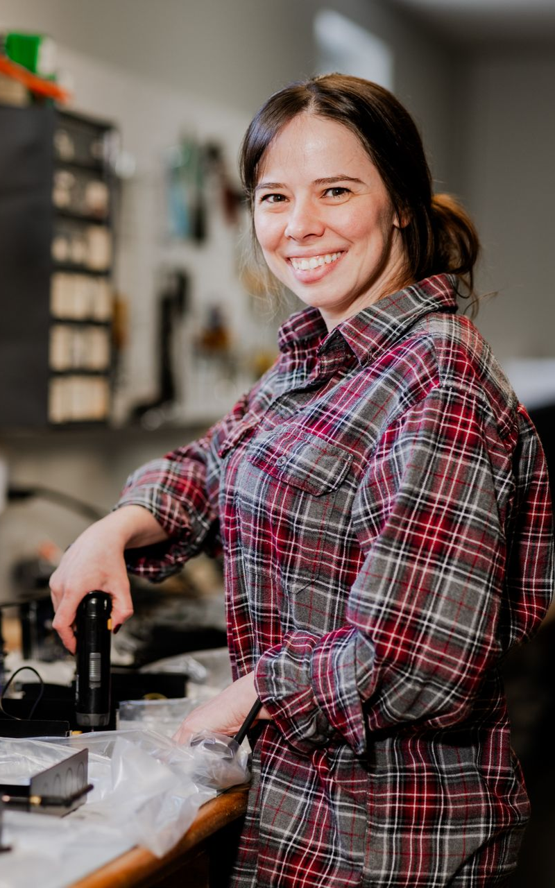 Woman smiling in a plaid shirt, holding a tool at a workbench in a workshop.