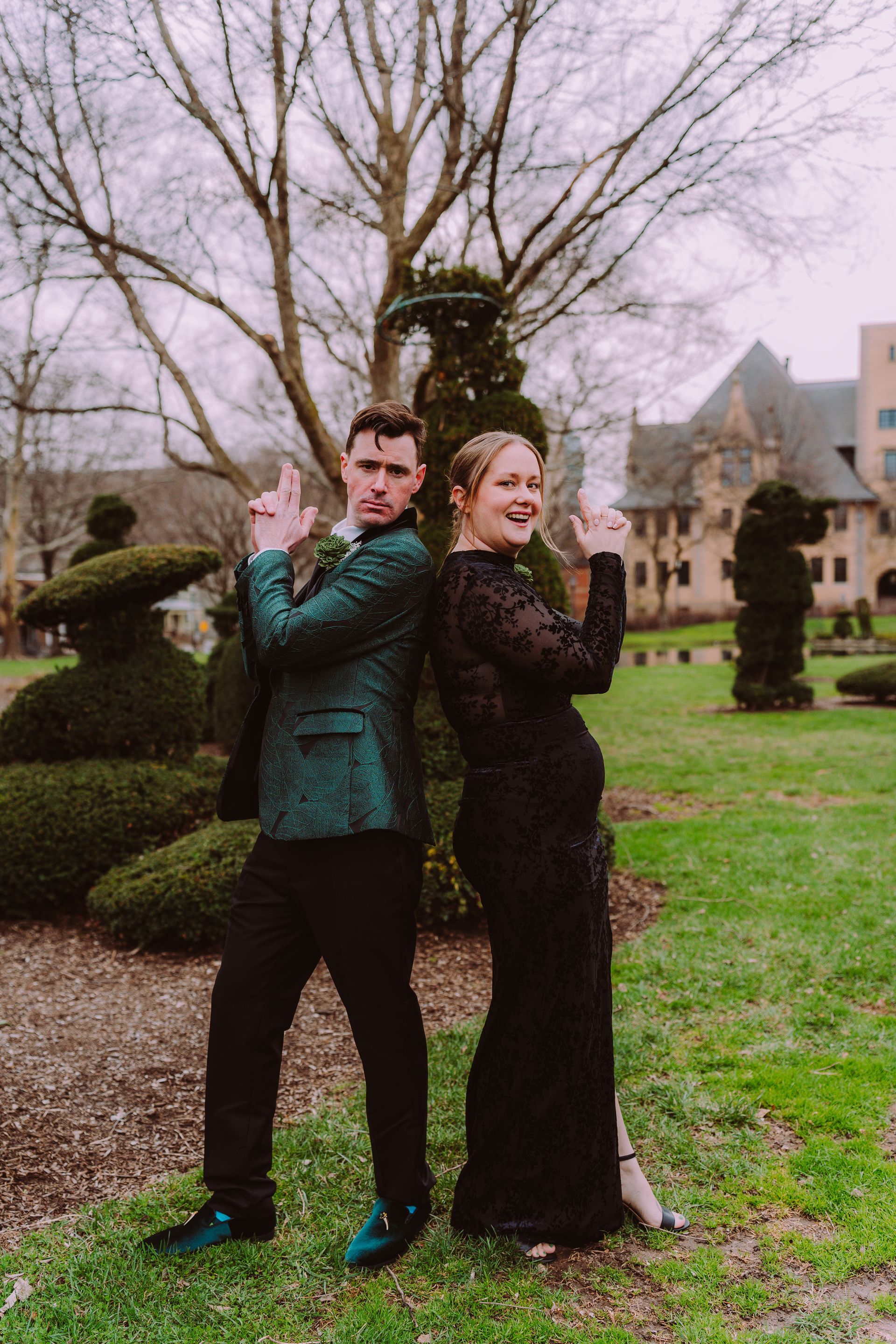 Two people in formal black and teal outfits posing back-to-back in a garden, clapping with smiles