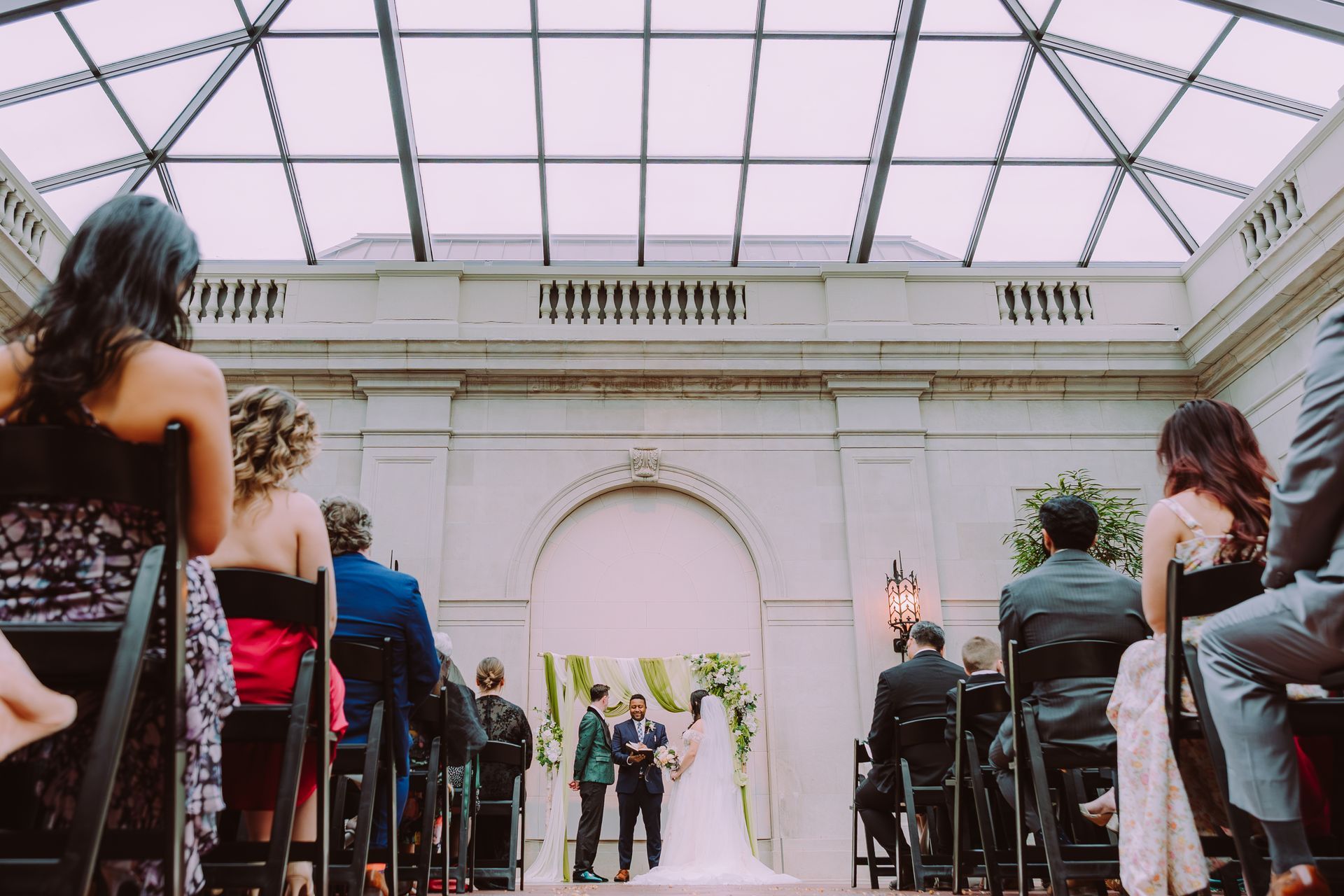 Guests seated in rows under a glass-roofed venue, watching a wedding ceremony at the altar