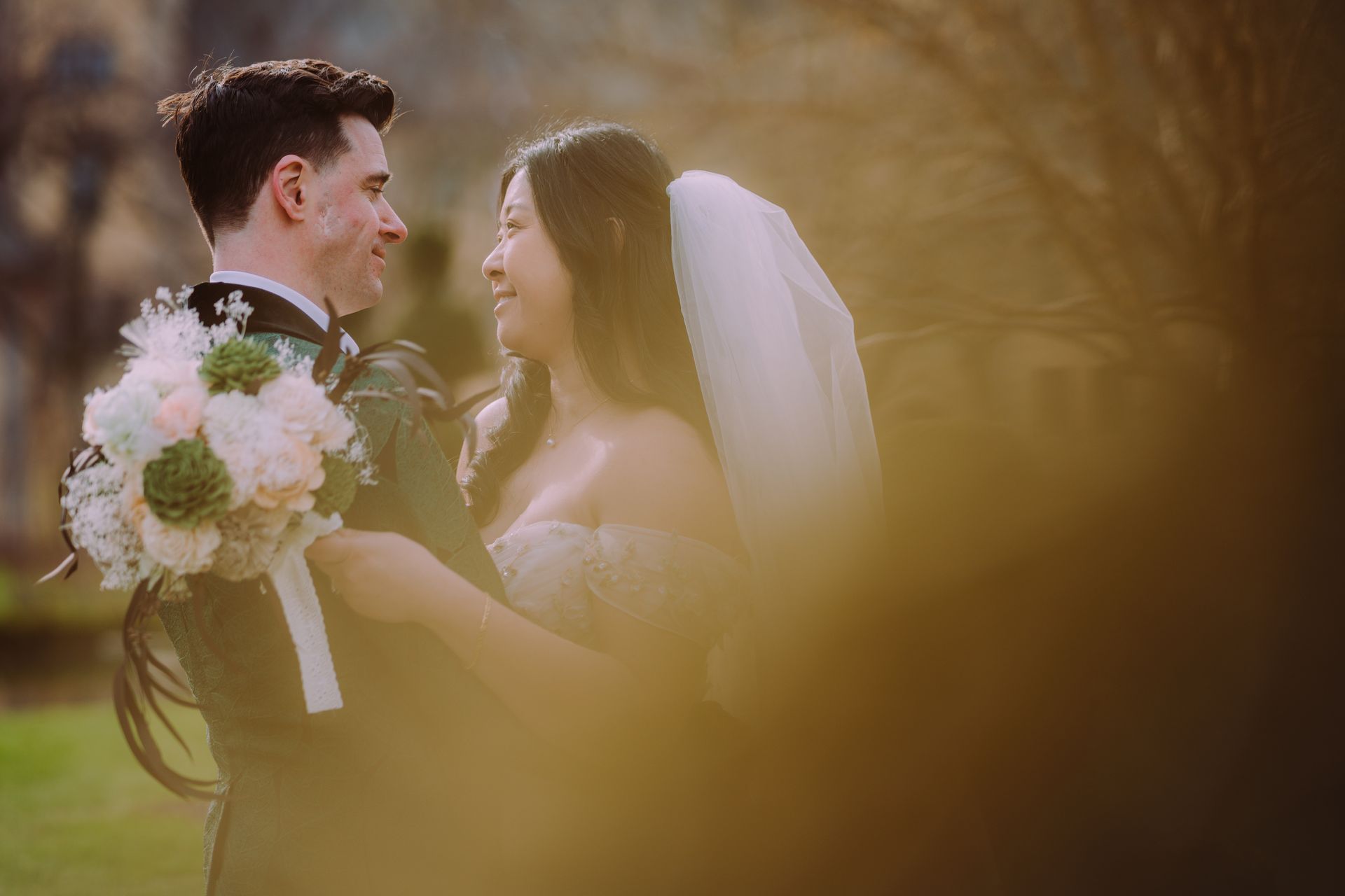 Bride and groom smiling in a sunlit garden, holding a bouquet during their wedding ceremony