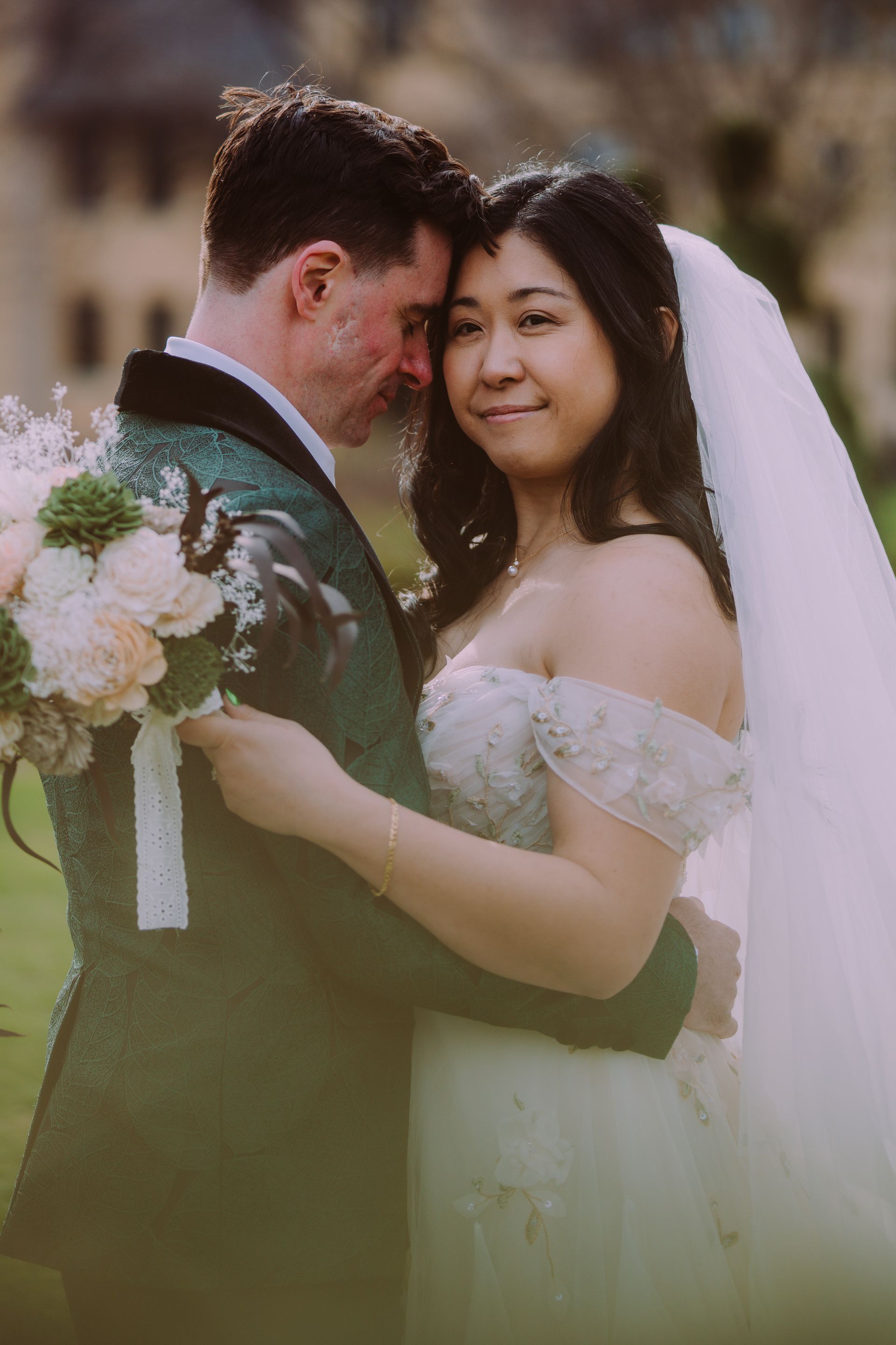 Wedding couple embracing outdoors, with bouquet and veil in soft sunlight