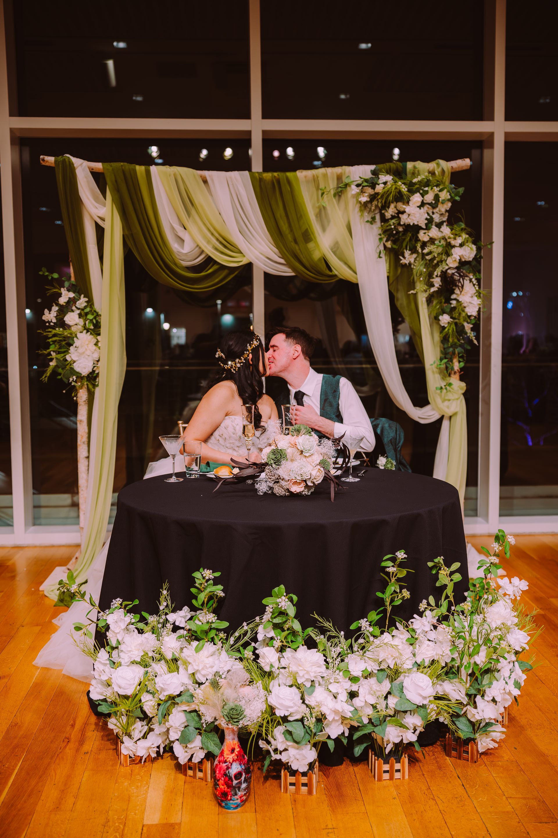 Couple kissing at a decorated wedding sweetheart table with white flowers and green drapery at night