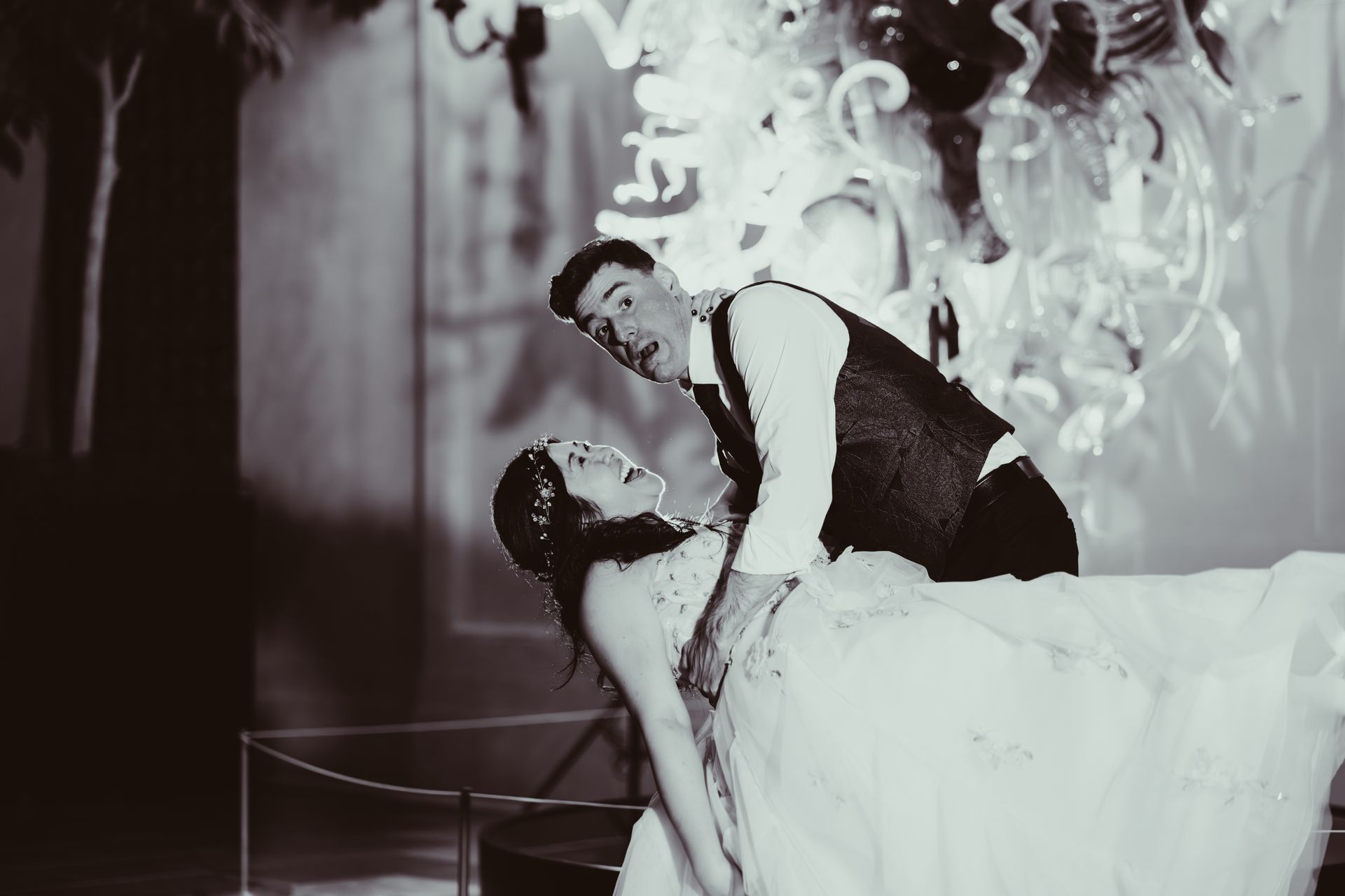 Two dancers in formal attire pose in a dim, elegant setting with dramatic black-and-white lighting.