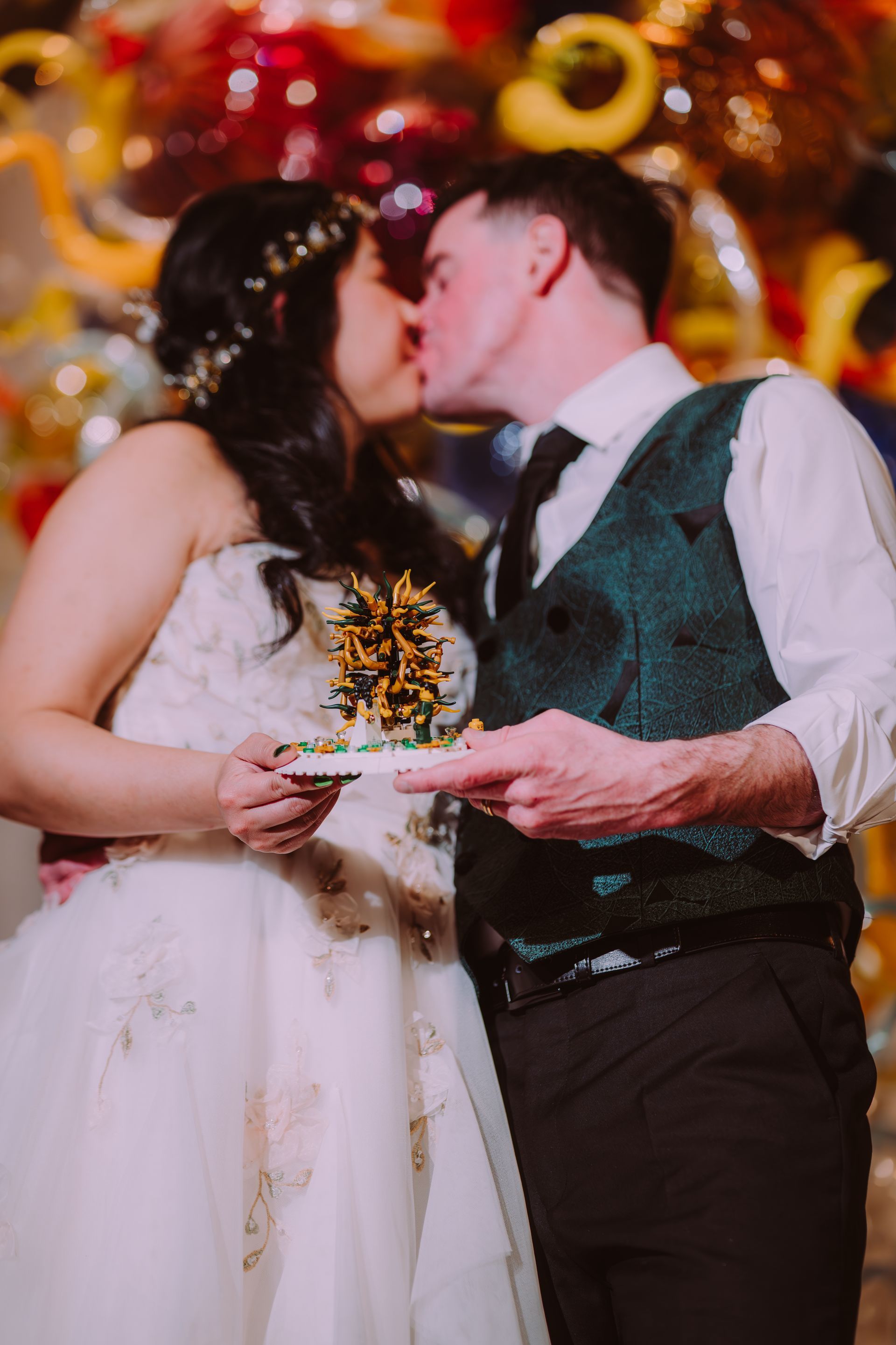 Bride and groom kiss on their wedding day, holding a bouquet amid colorful balloons.