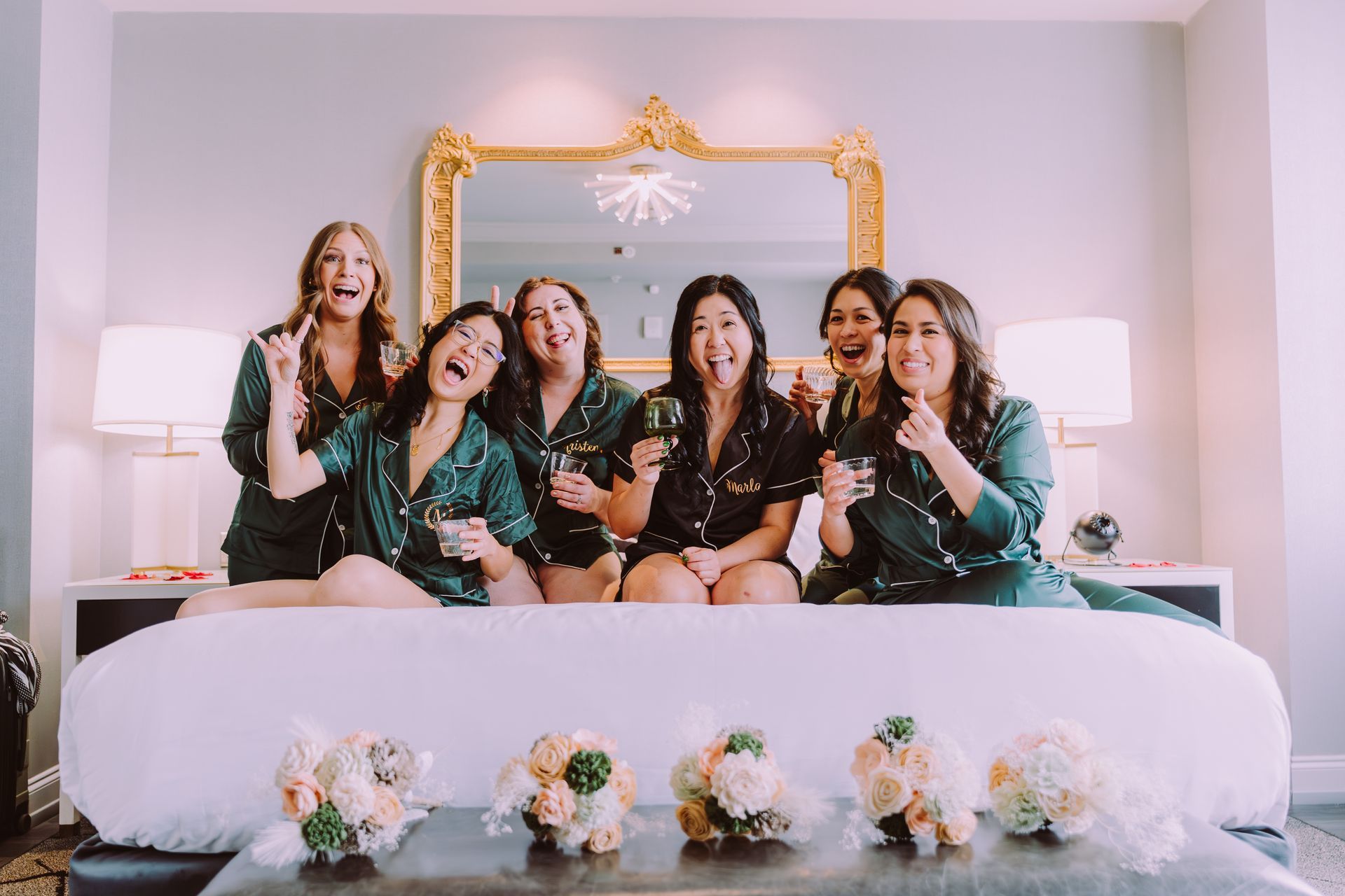 Five women in green pajamas sitting on a bed in a decorated bedroom, smiling and posing together