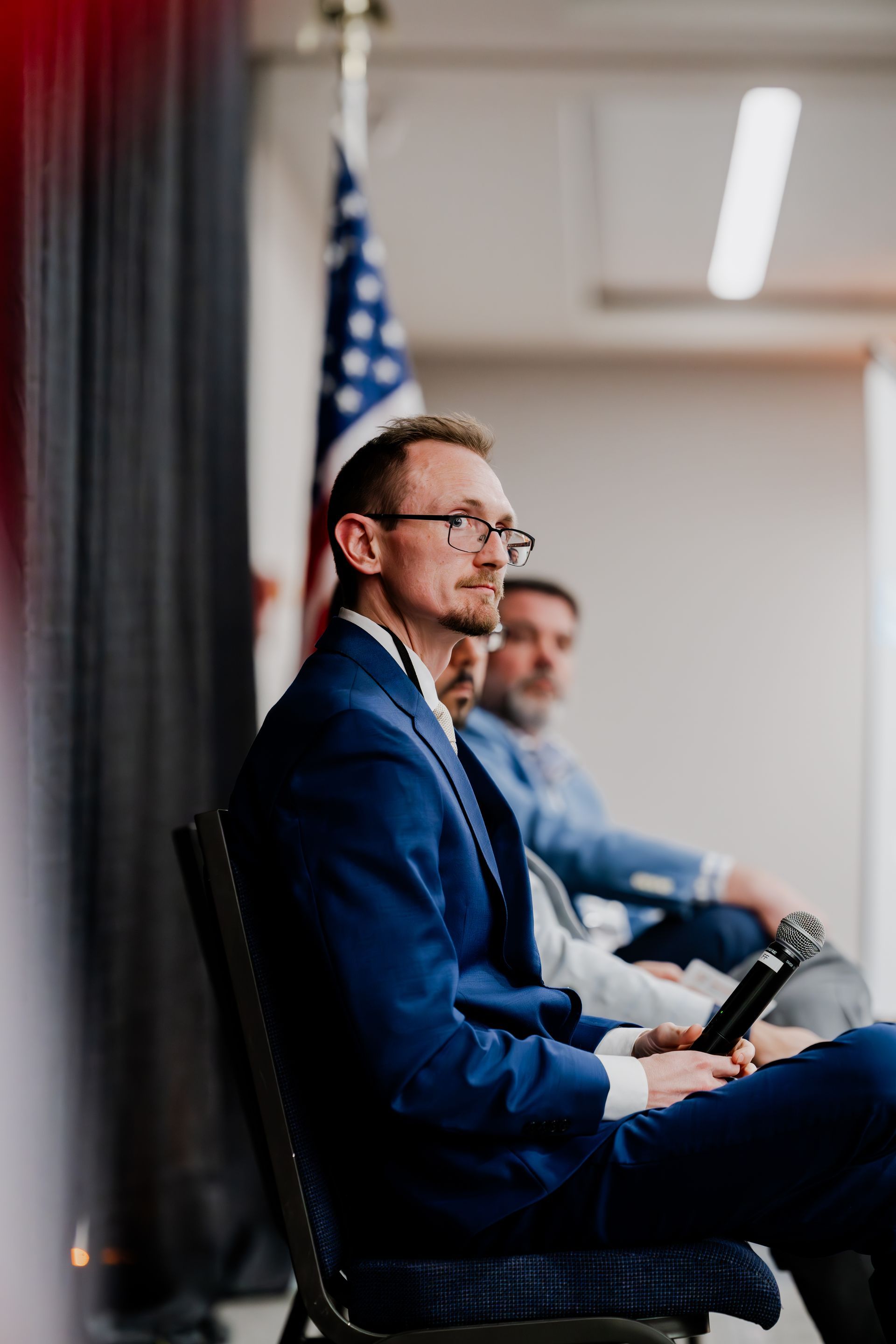 A man in a suit is sitting in a chair with a microphone in front of an american flag.