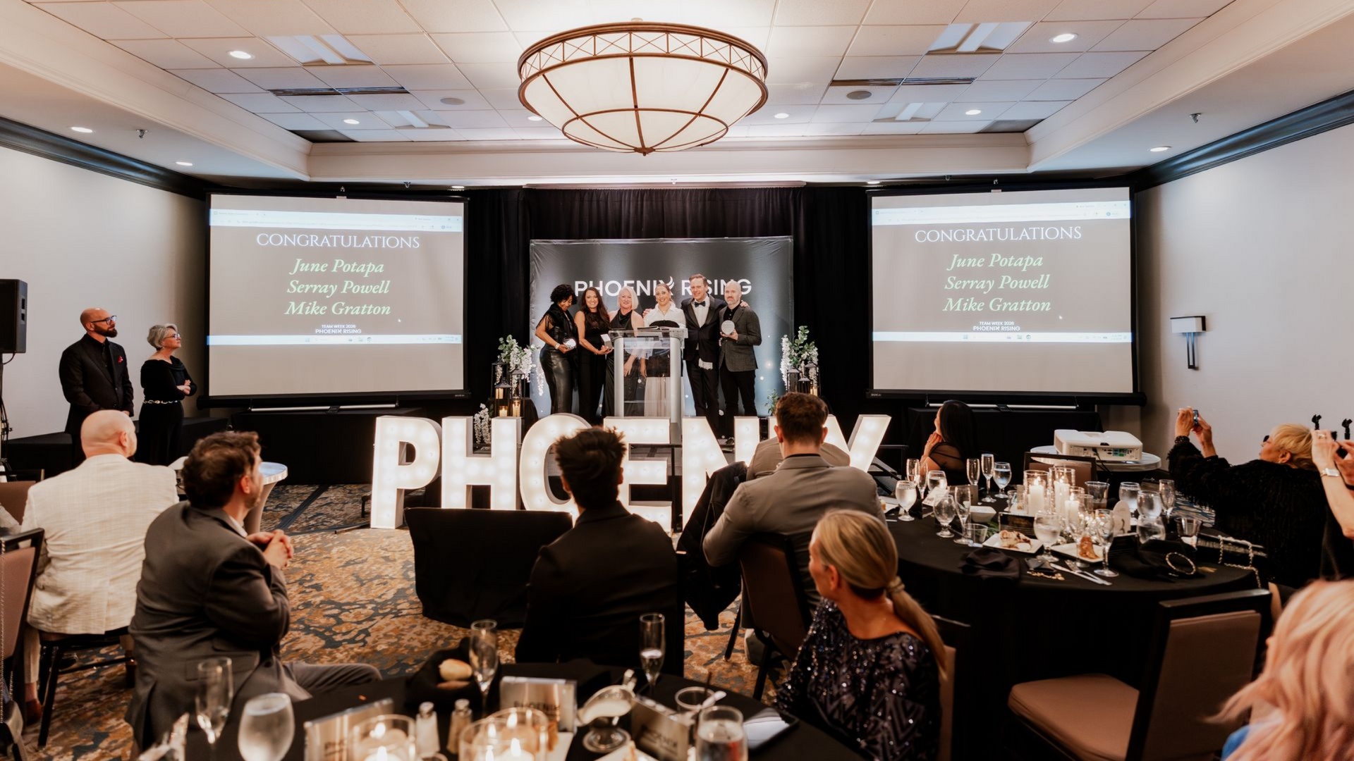 A group of people standing on a stage at an awards ceremony with giant illuminated "PHOENIX" letters in the foreground.