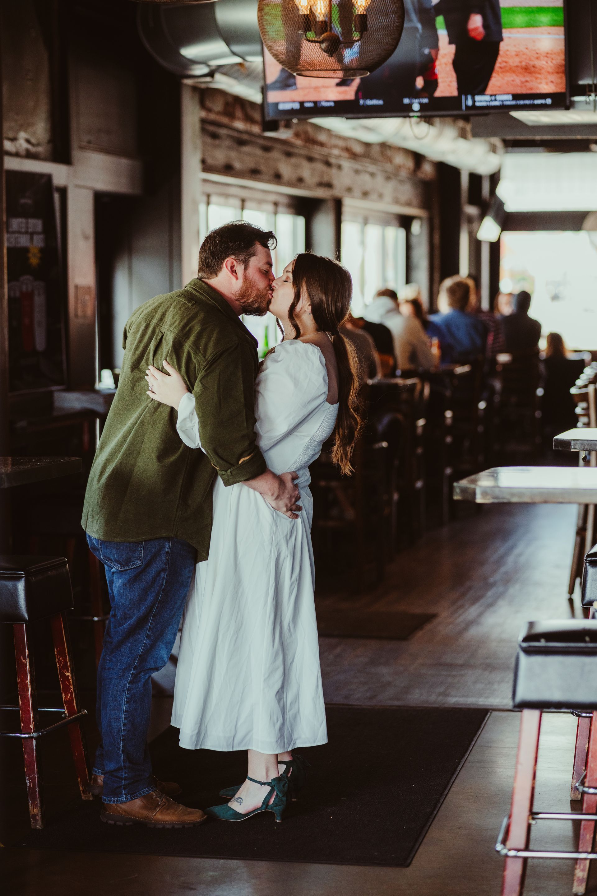 Couple kissing in a dimly lit bar with a TV overhead and stools along the counter