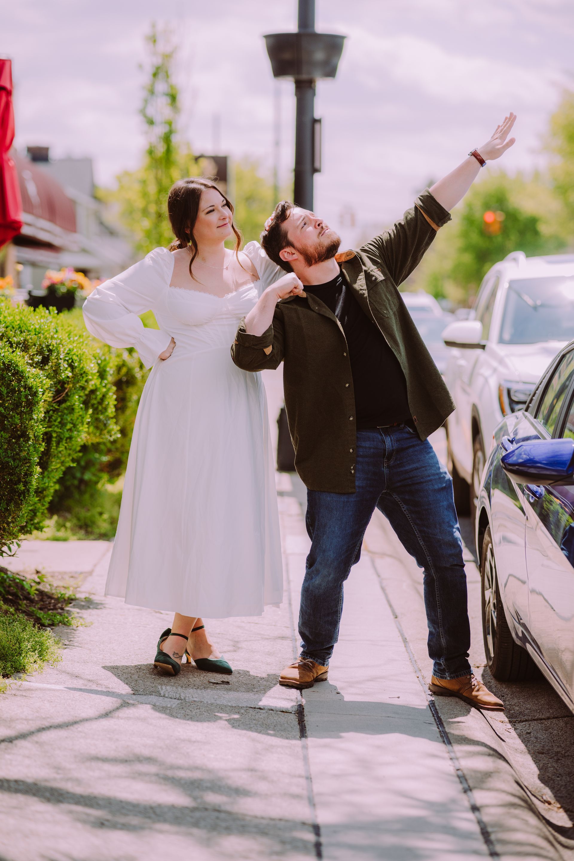 Two people posing on a sunlit sidewalk, smiling and gesturing playfully beside parked cars.