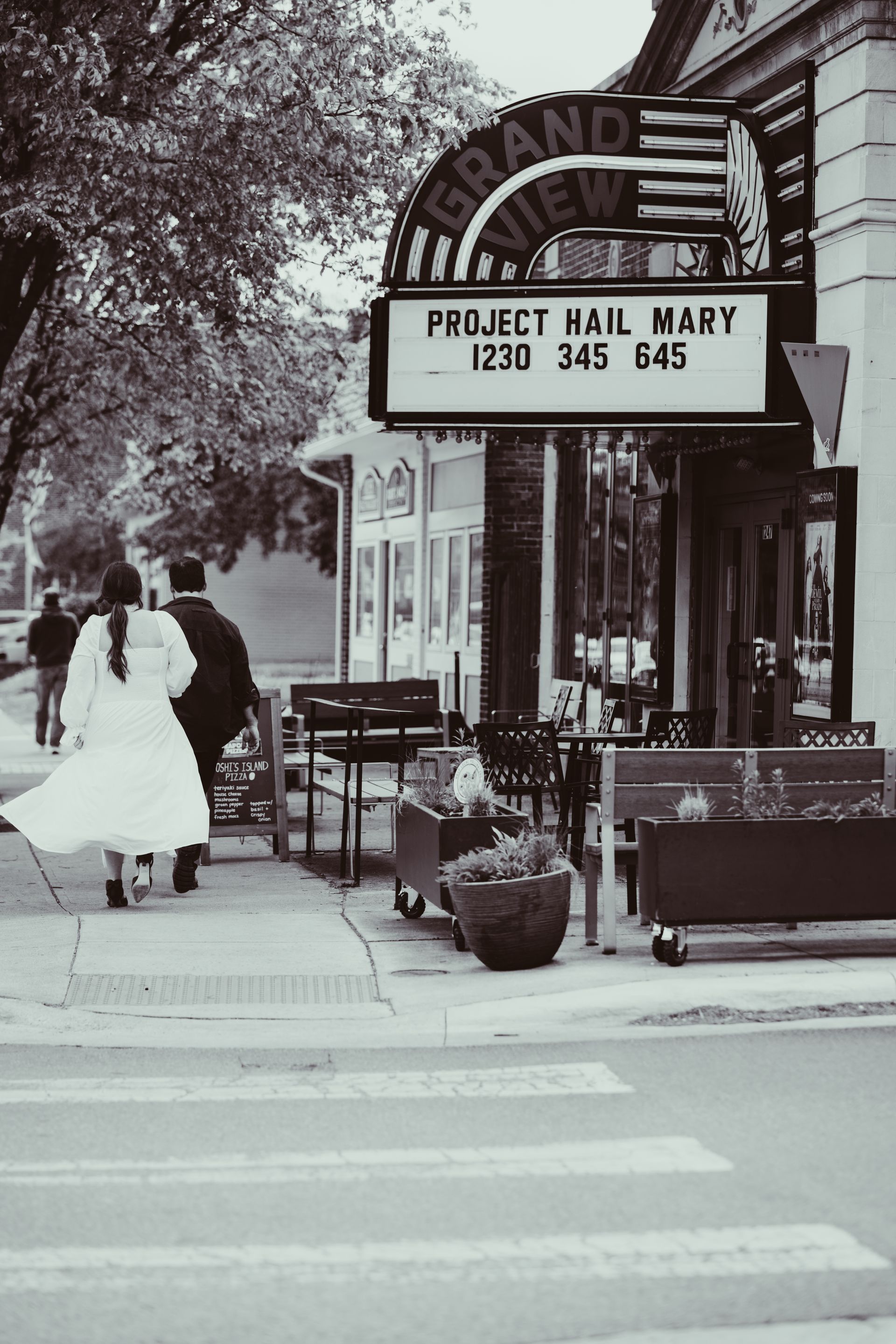 Two people walking past a storefront with a “Project Hall” sign on a city sidewalk.