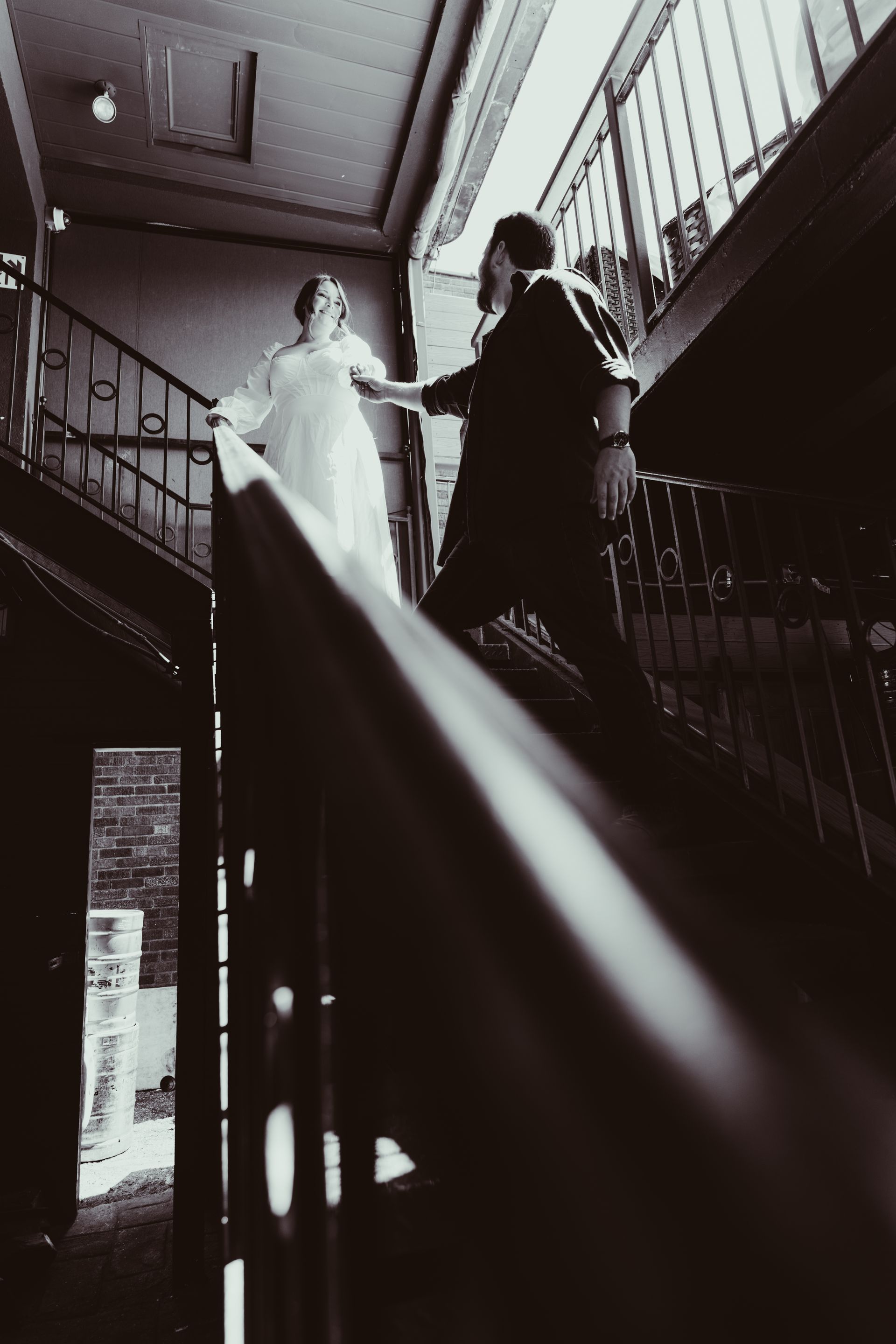 Two people stand on a staircase, one in a white dress and one in dark clothing, with bright light streaming in.
