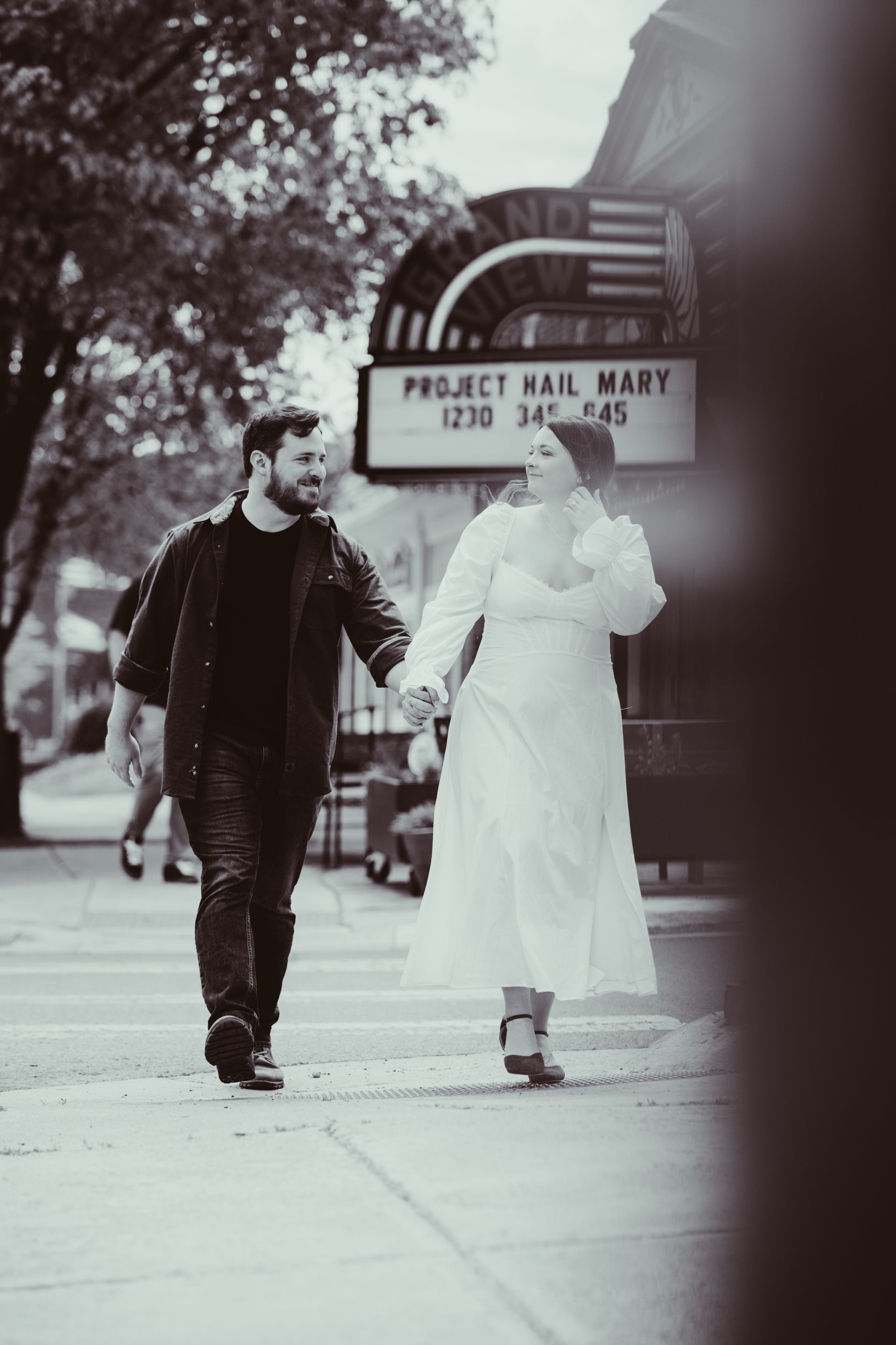Two people walking on a sidewalk past a Project Hall Market sign, one in dark clothes and one in a light coat