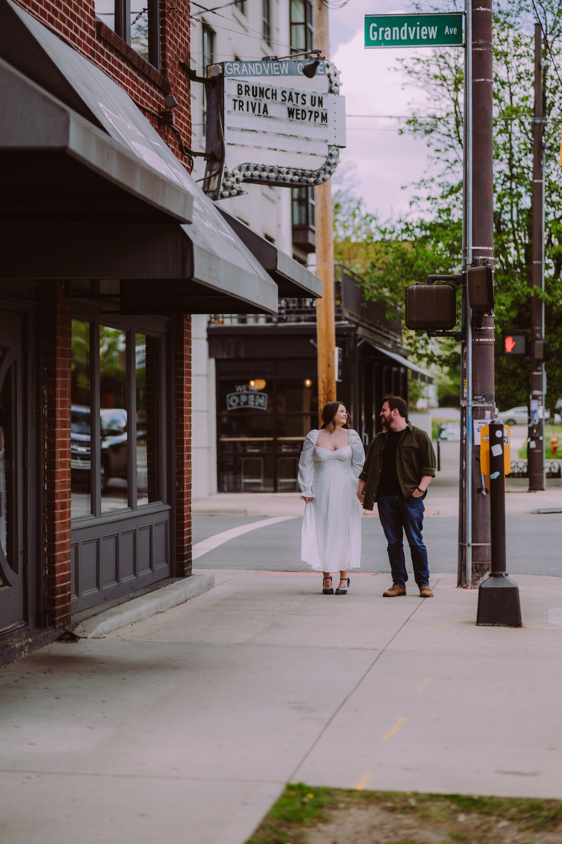 Two people walking on a sidewalk beside storefronts and a street sign in a city scene