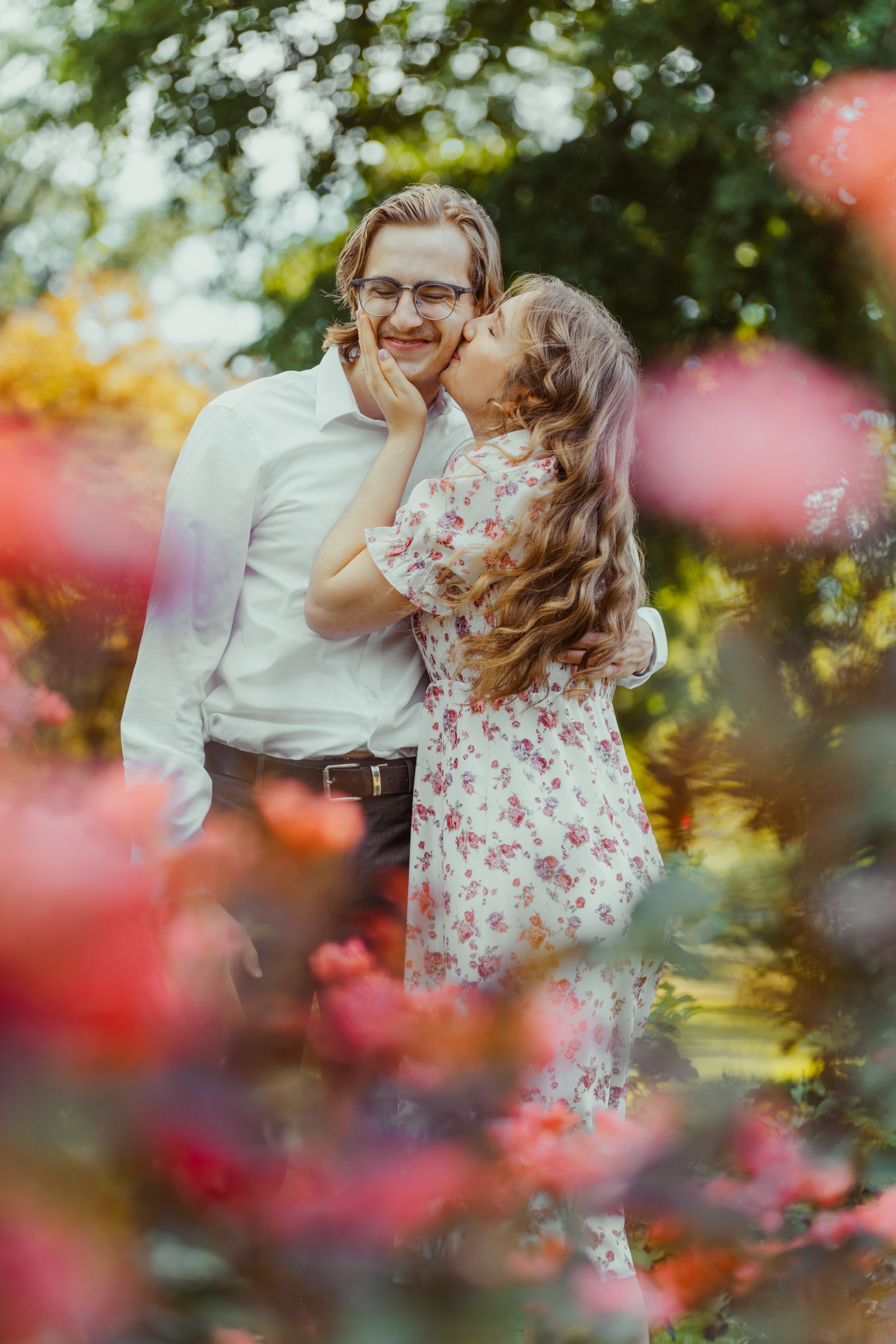 A woman is kissing a man on the cheek in a park surrounded by flowers.