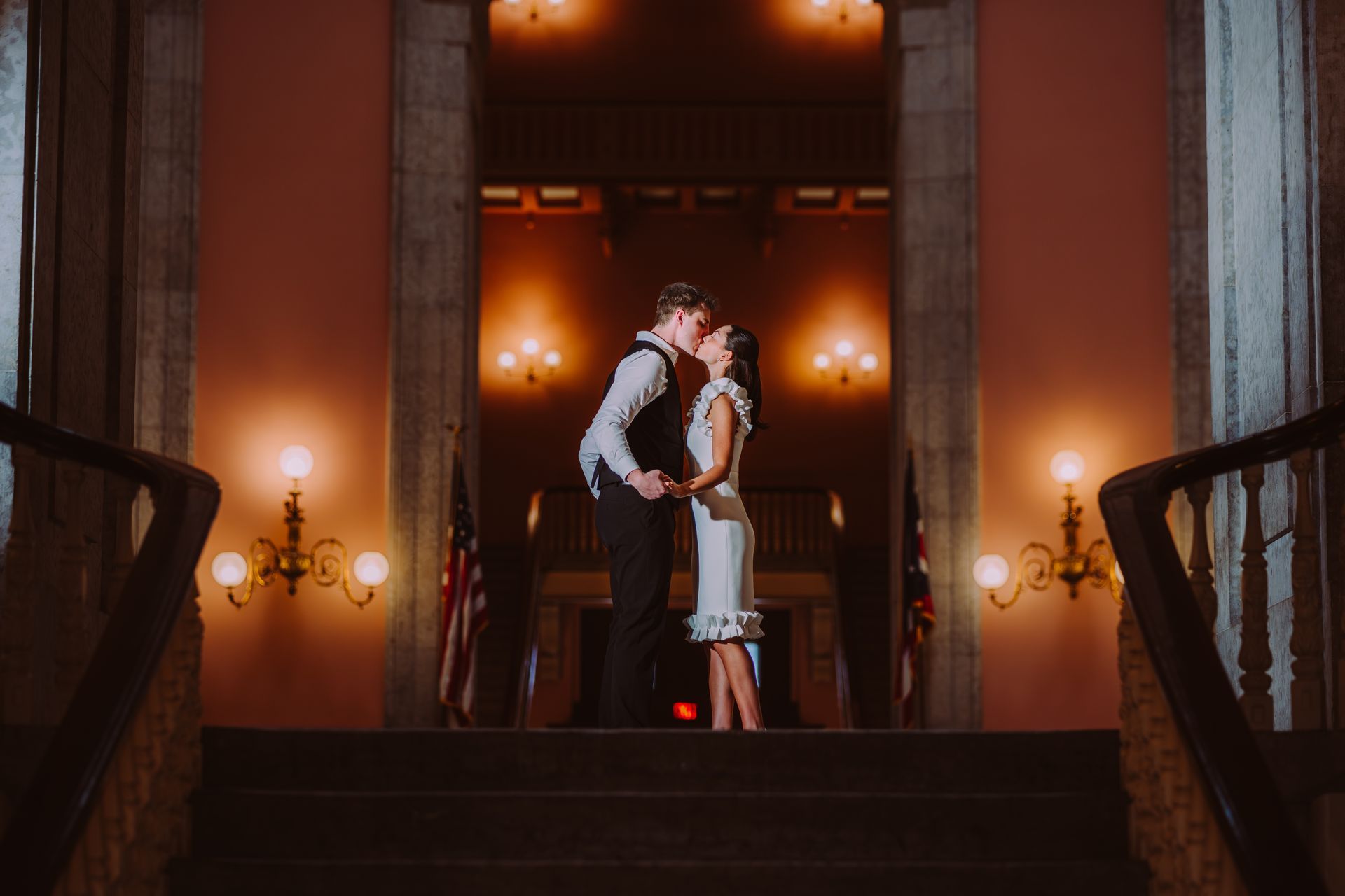 Couple standing on a grand staircase in a warmly lit, elegant hall, about to kiss