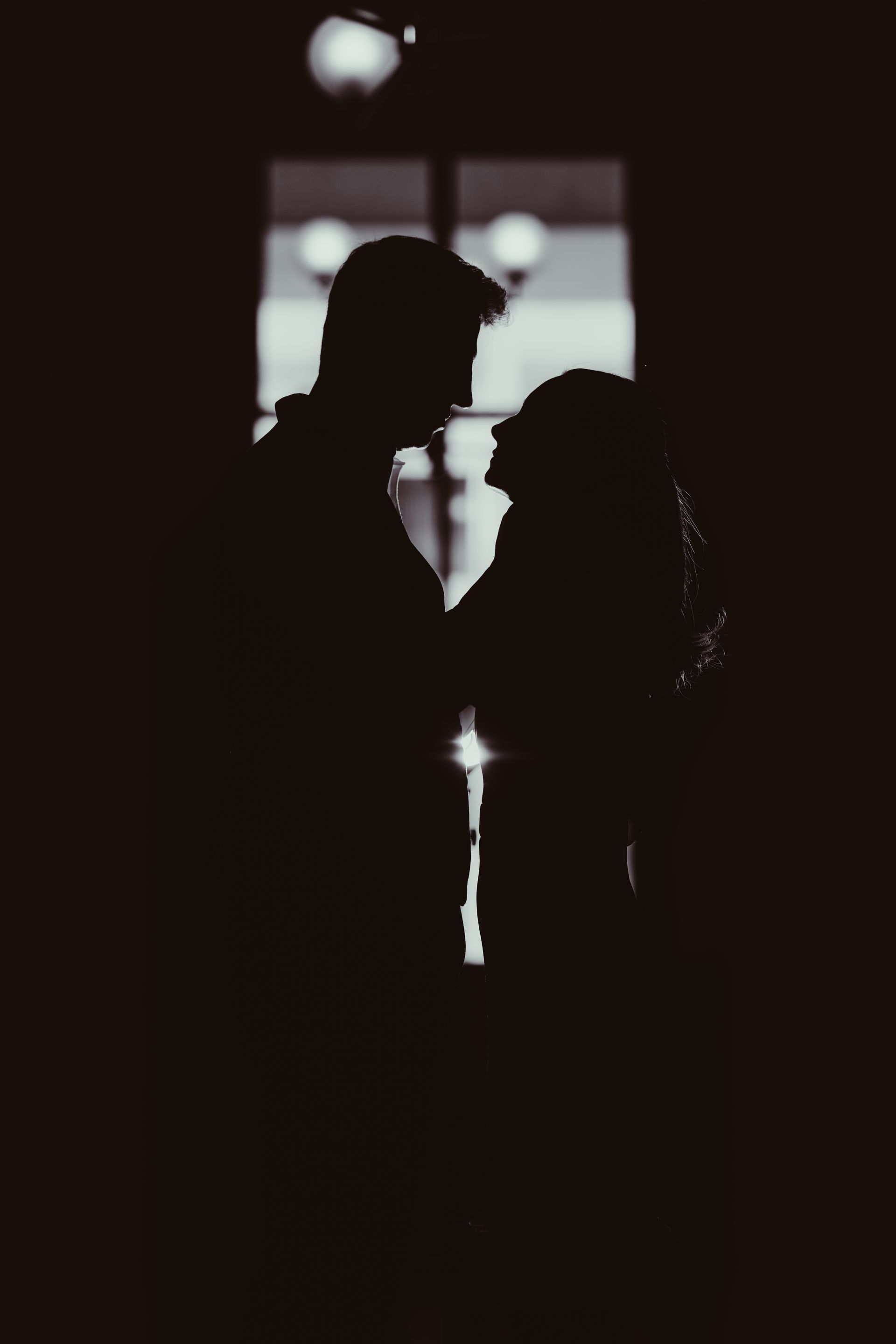 Silhouetted couple facing each other in a dimly lit hallway, backlit by a bright doorway