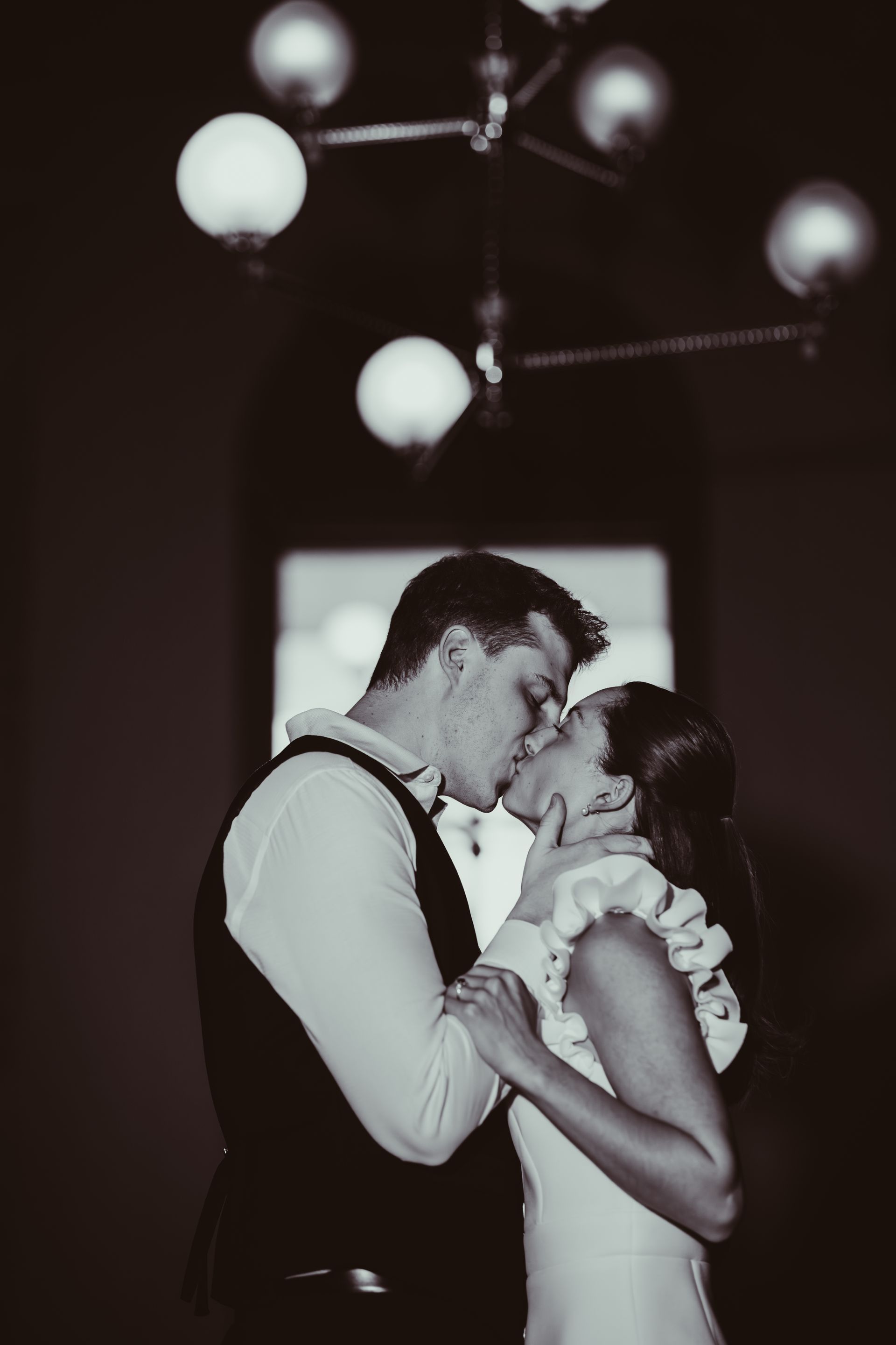 A couple kisses during their wedding, illuminated by a decorative chandelier in a dark room.