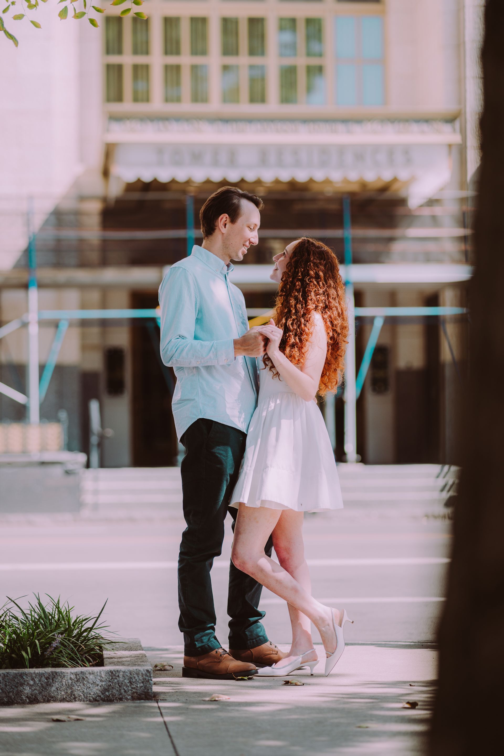 Couple facing each other and smiling. Woman in white dress, man in blue shirt, in front of a building.