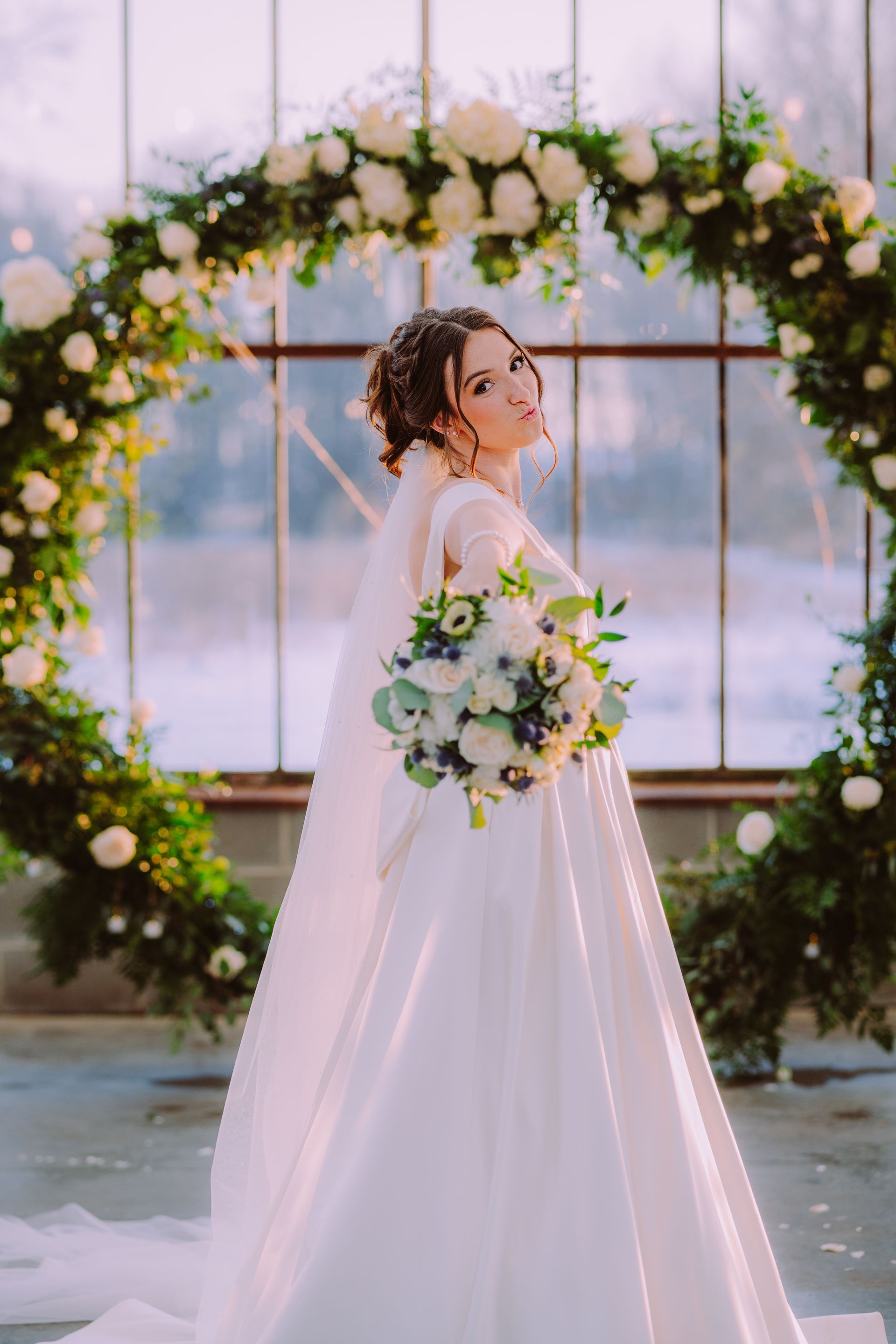 Bride in white gown holding bouquet, standing before floral arch in greenhouse.