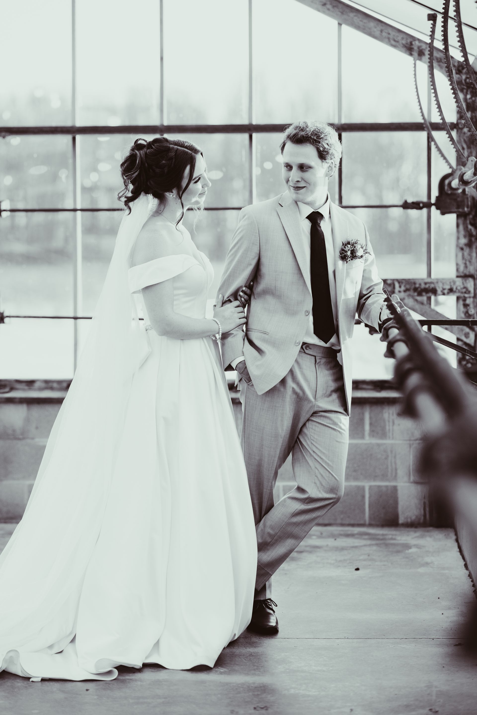 Bride and groom in wedding attire smiling at each other in a greenhouse.