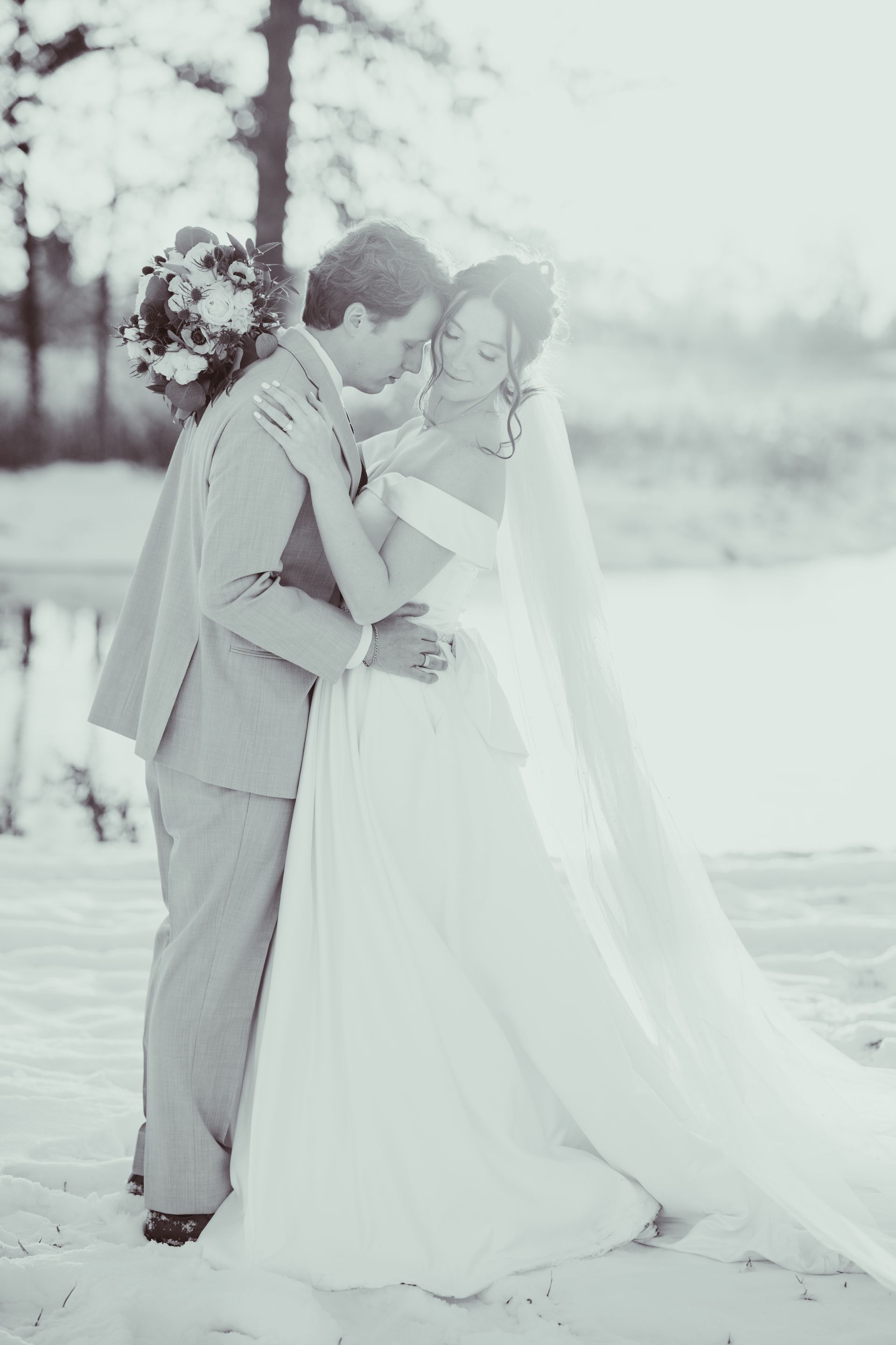 Bride and groom embrace on a beach. Bride in a flowing gown, groom in a suit. Bright sunlight.