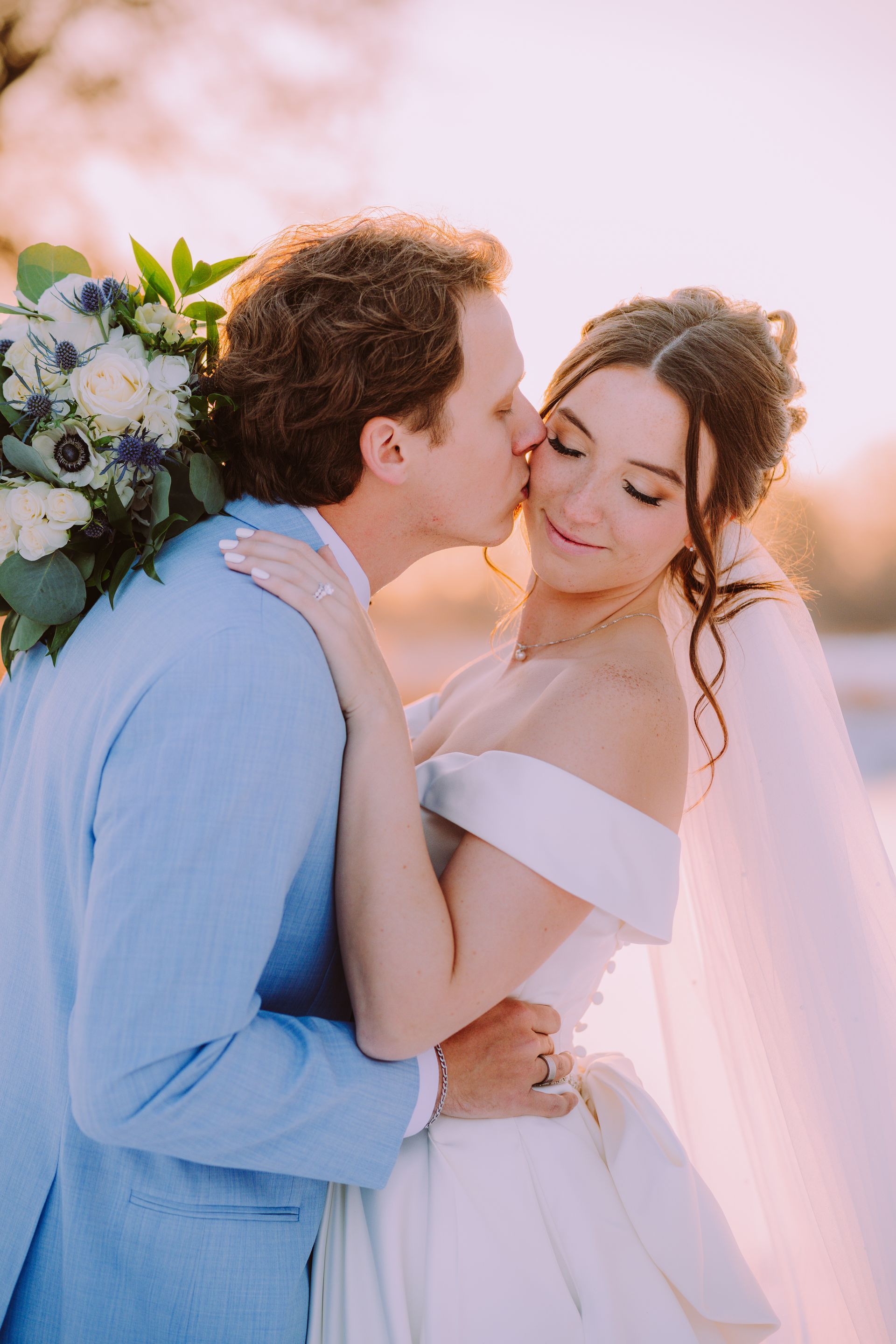 Groom kissing bride's cheek; couple embraces outdoors. Bride wears off-shoulder gown, veil.
