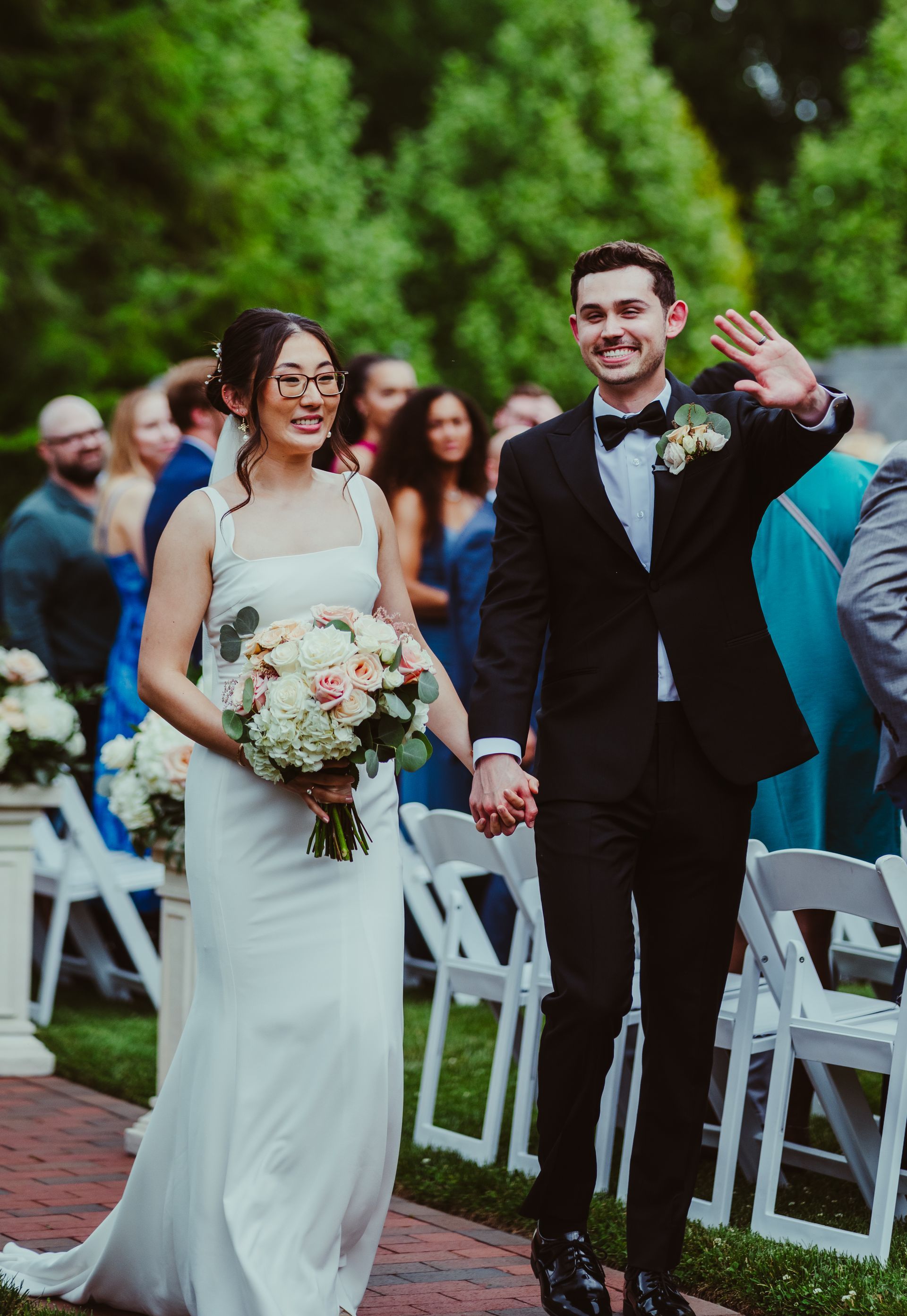 Bride and groom walking down an outdoor aisle, smiling and waving among seated wedding guests