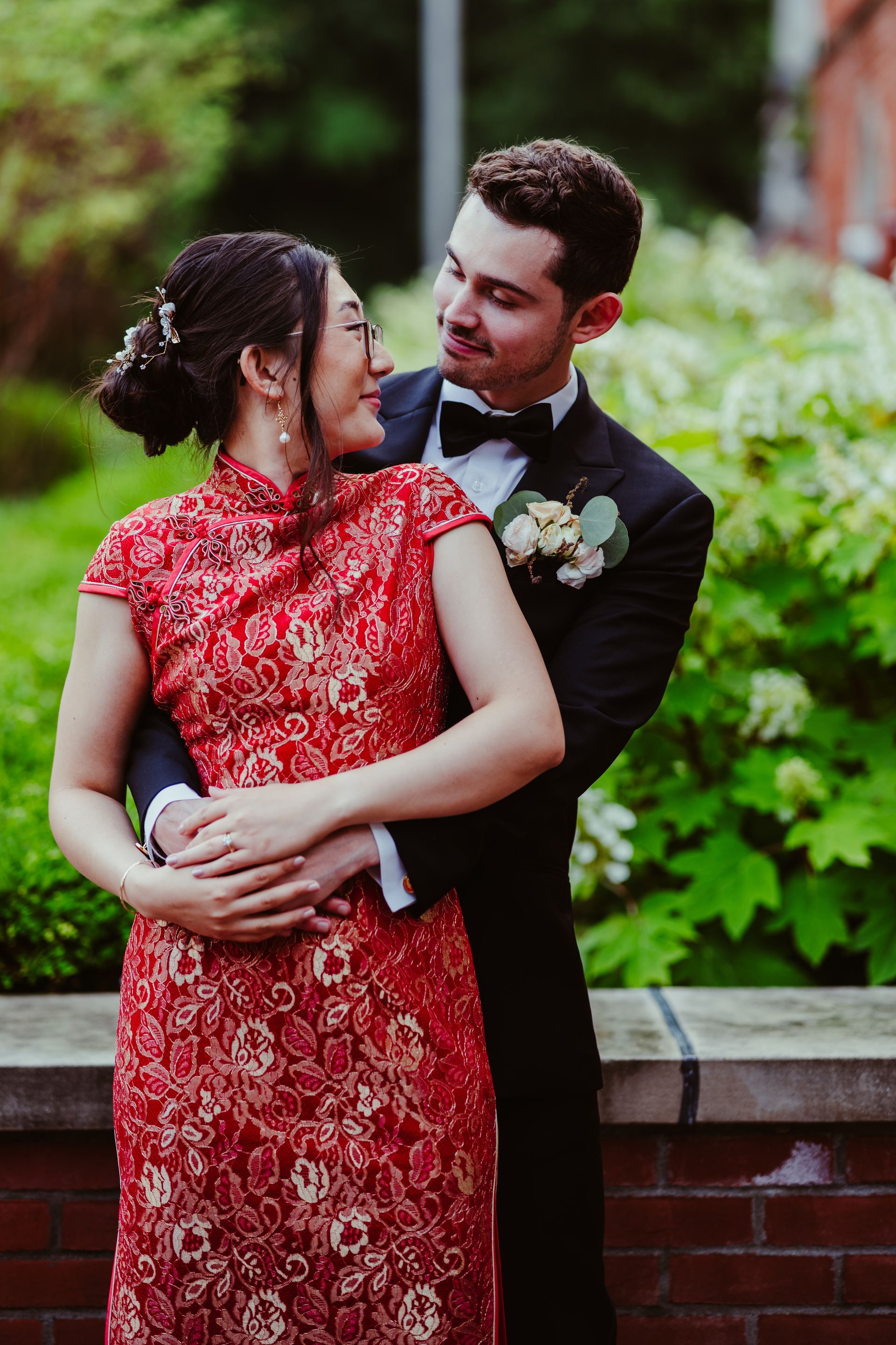 Couple in formal attire embracing outdoors, with red dress, black suit, and green foliage backdrop
