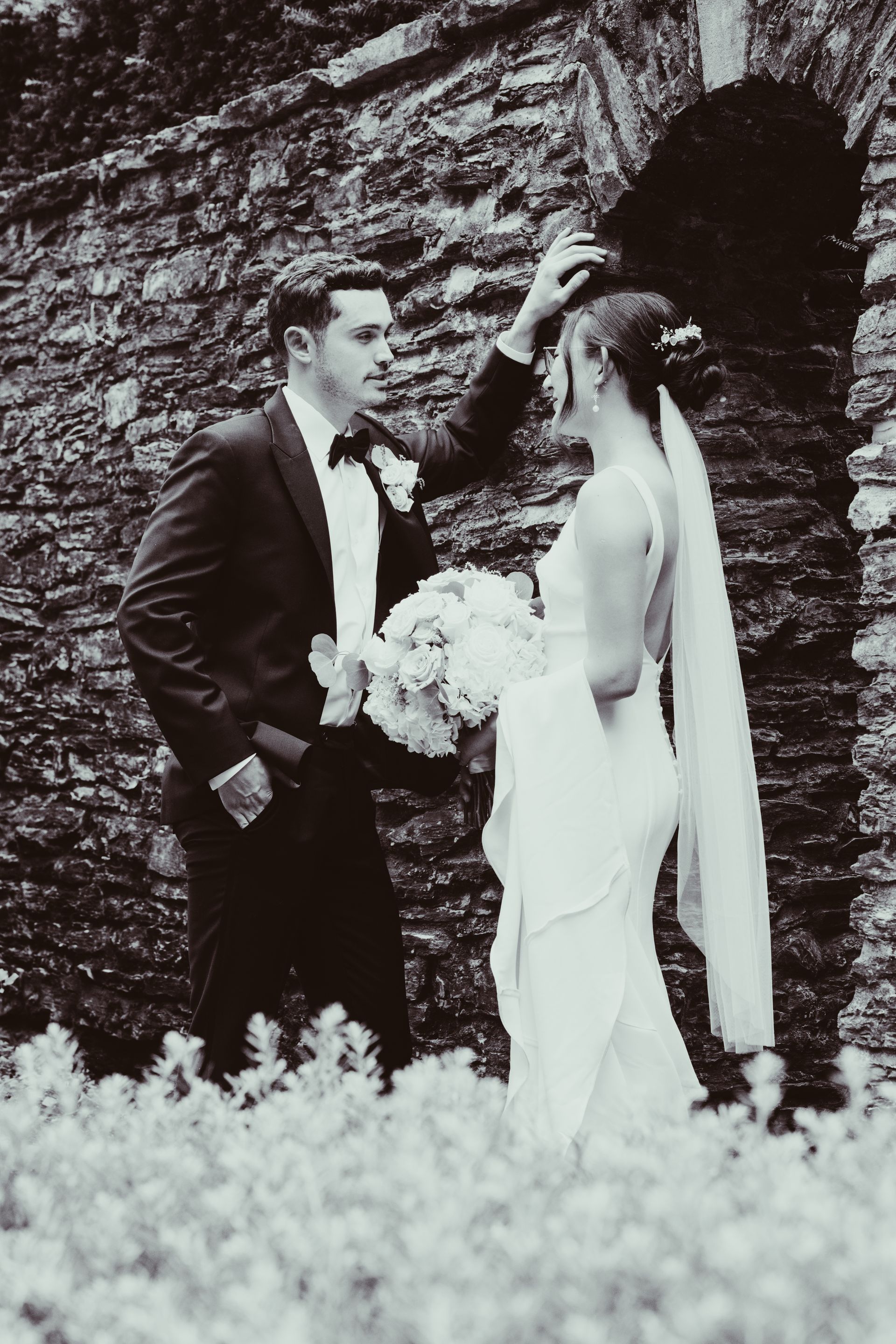 Bride and groom pose in formal attire beside a rocky wall, holding a bouquet outdoors.