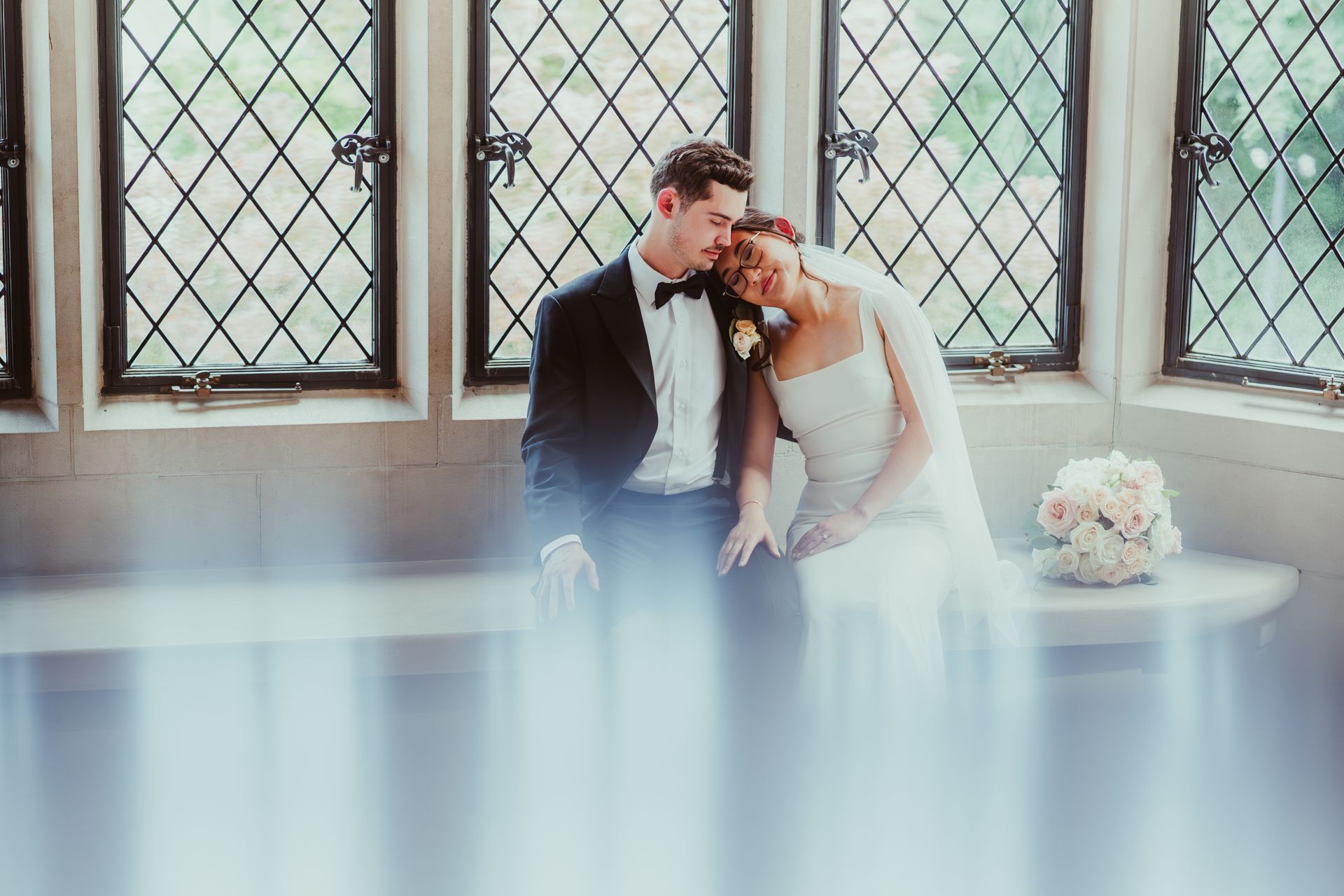 Bride and groom seated by windows, leaning together in a soft-lit wedding portrait