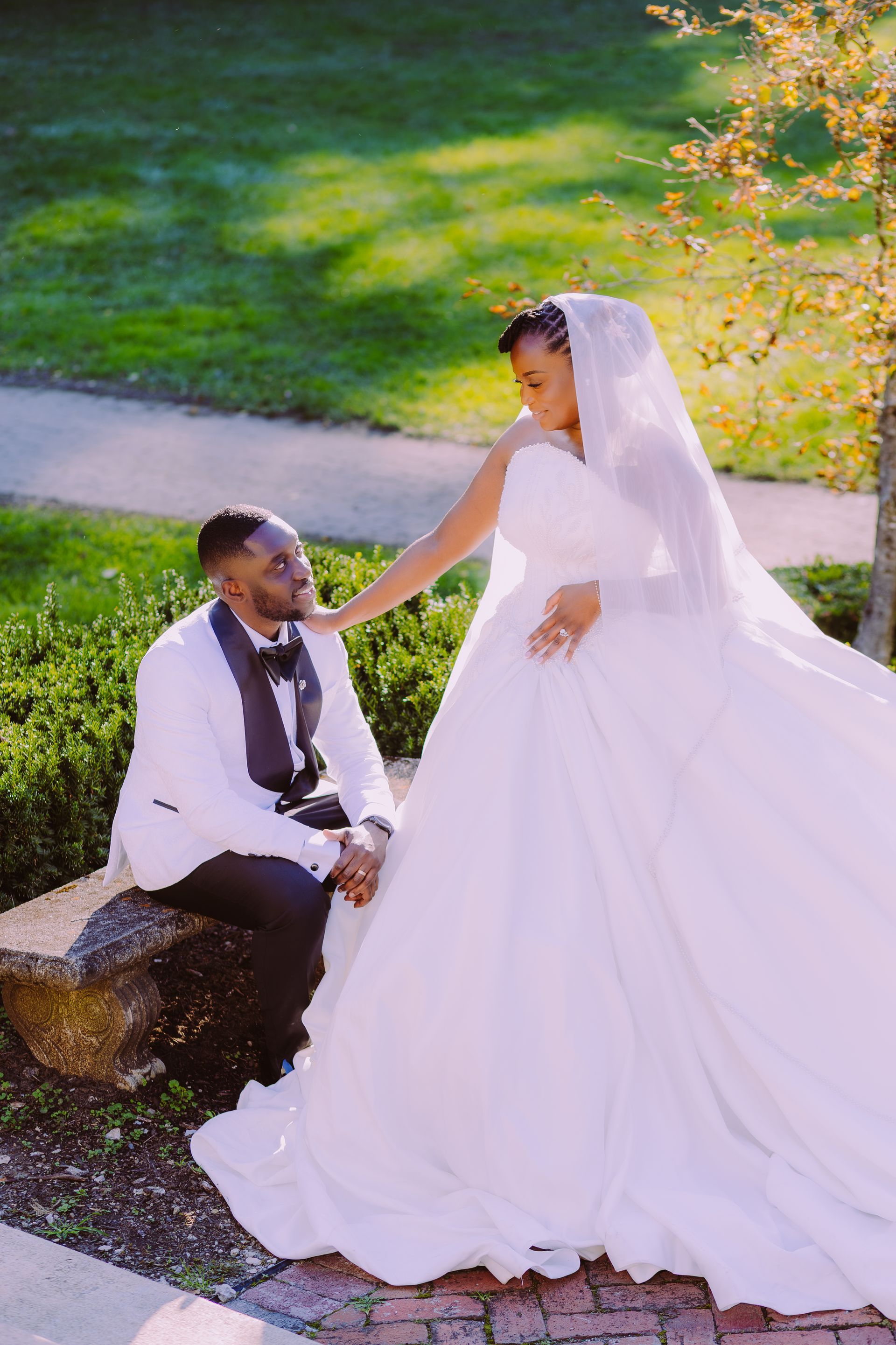 Bride in white gown and veil standing beside groom seated outdoors on a stone bench.