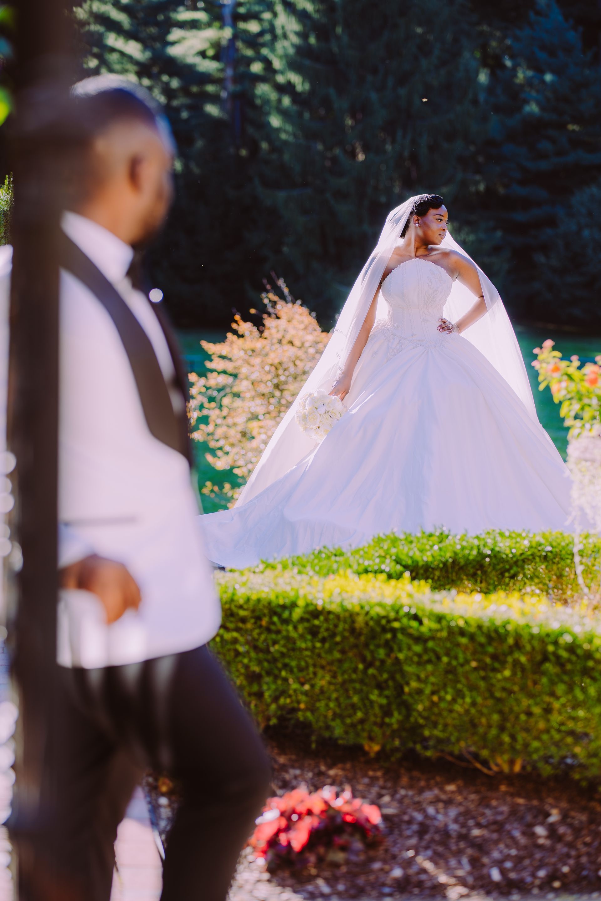 Bride in a white gown and veil standing outdoors behind a blurred foreground figure, surrounded by flowers and greenery