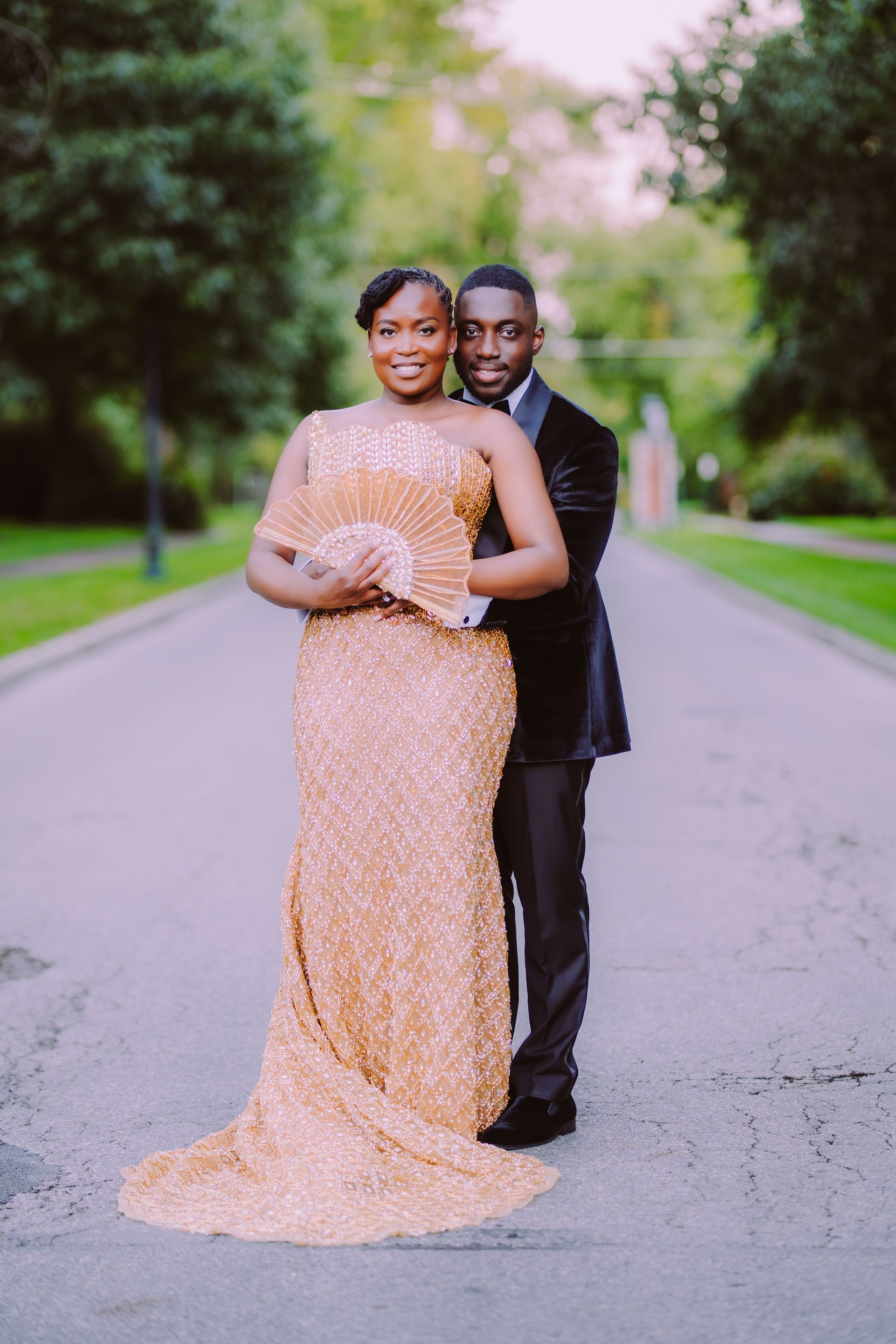 Couple posing on a tree-lined road, woman in a sparkling peach gown and man in a black suit.