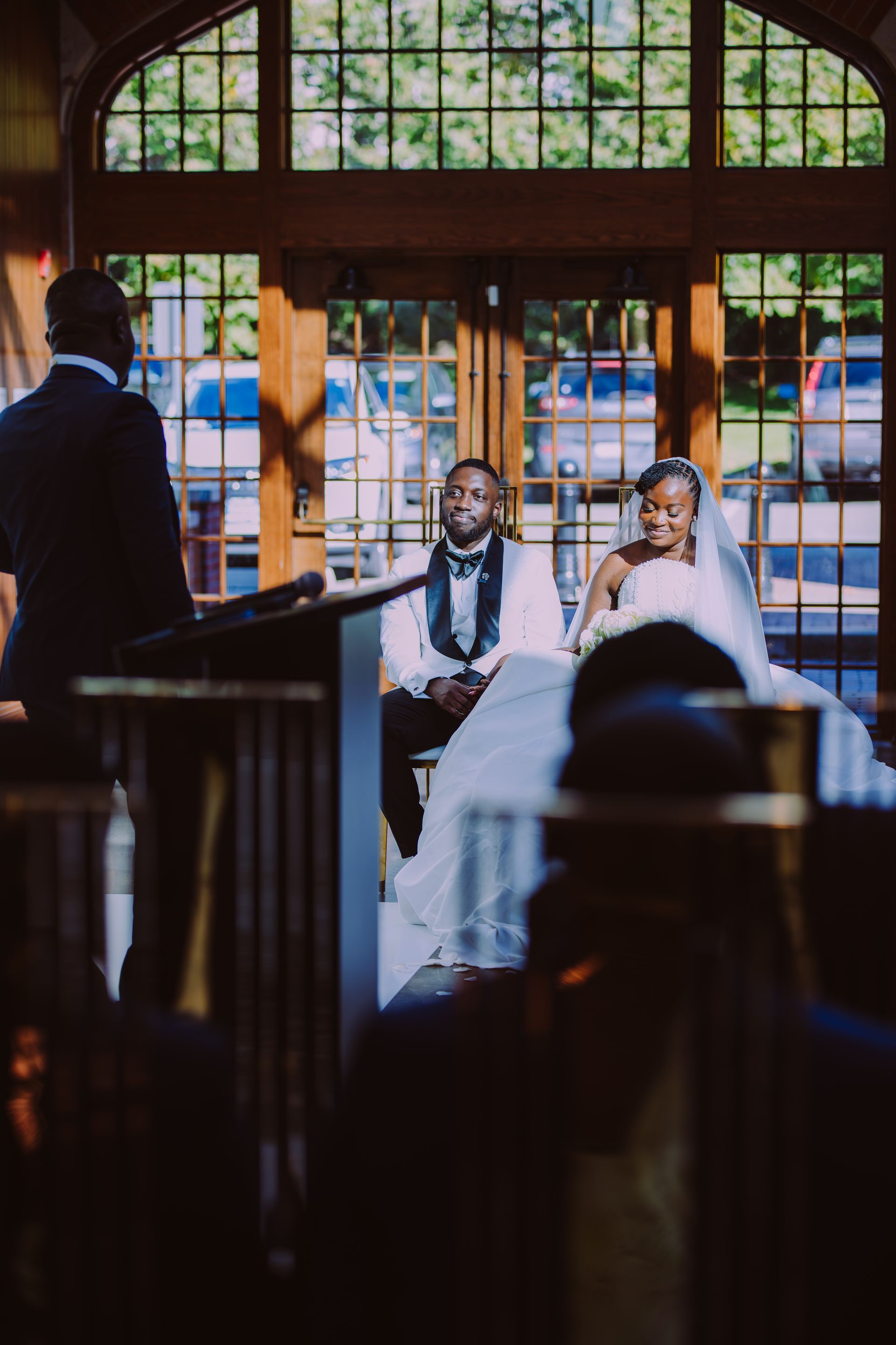 Bride and groom seated at altar in a church, viewed from the aisle with stained glass behind them