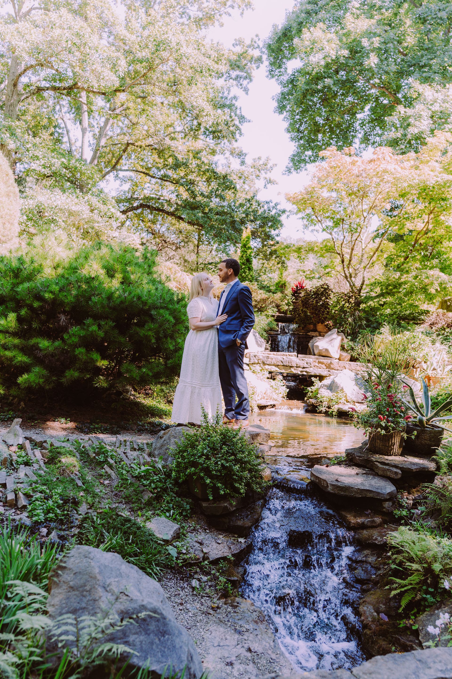 Couple in formal attire standing by a small waterfall in a lush garden, surrounded by trees and rocks