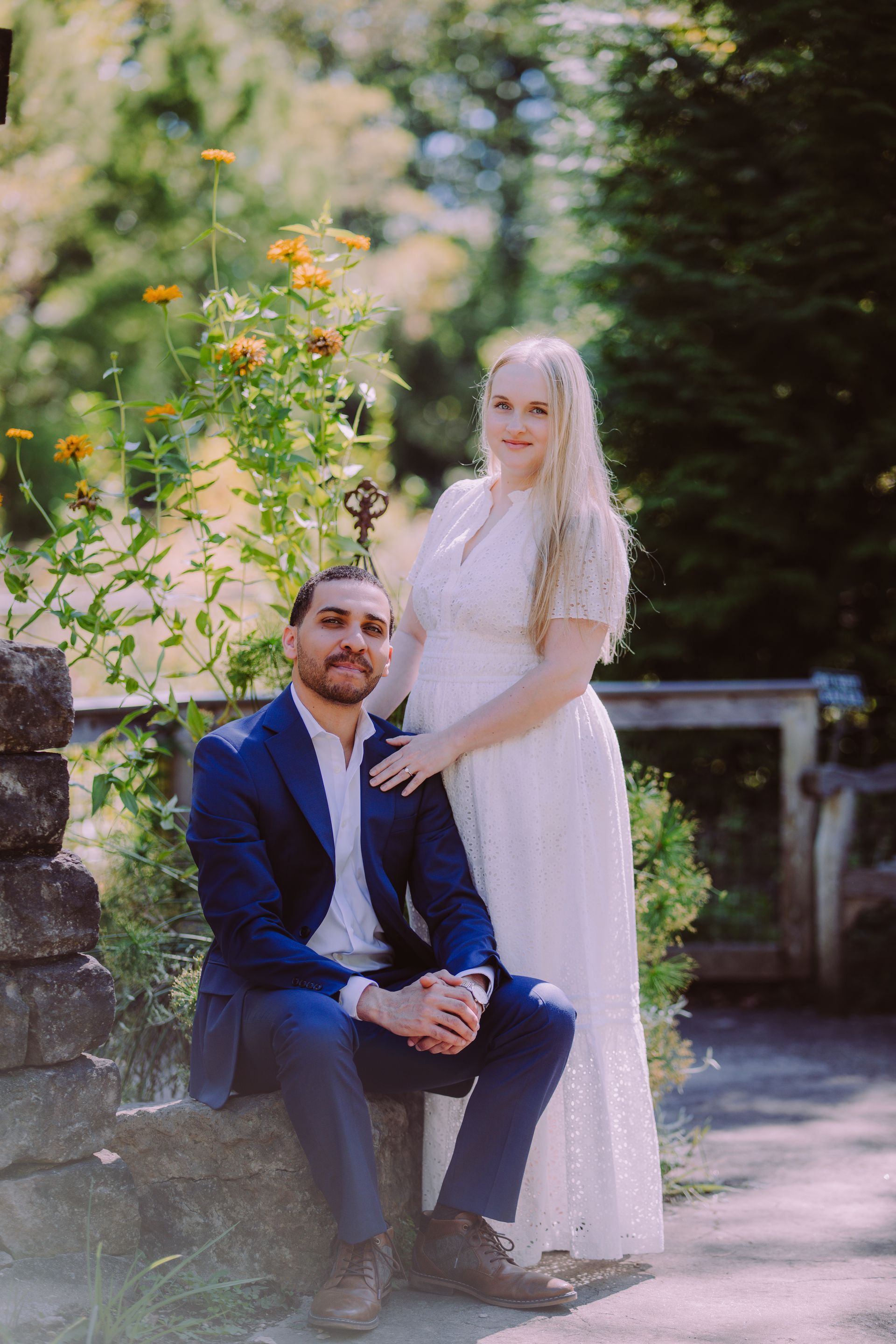Couple posing outdoors in wedding attire, with the man seated and the woman standing beside him.