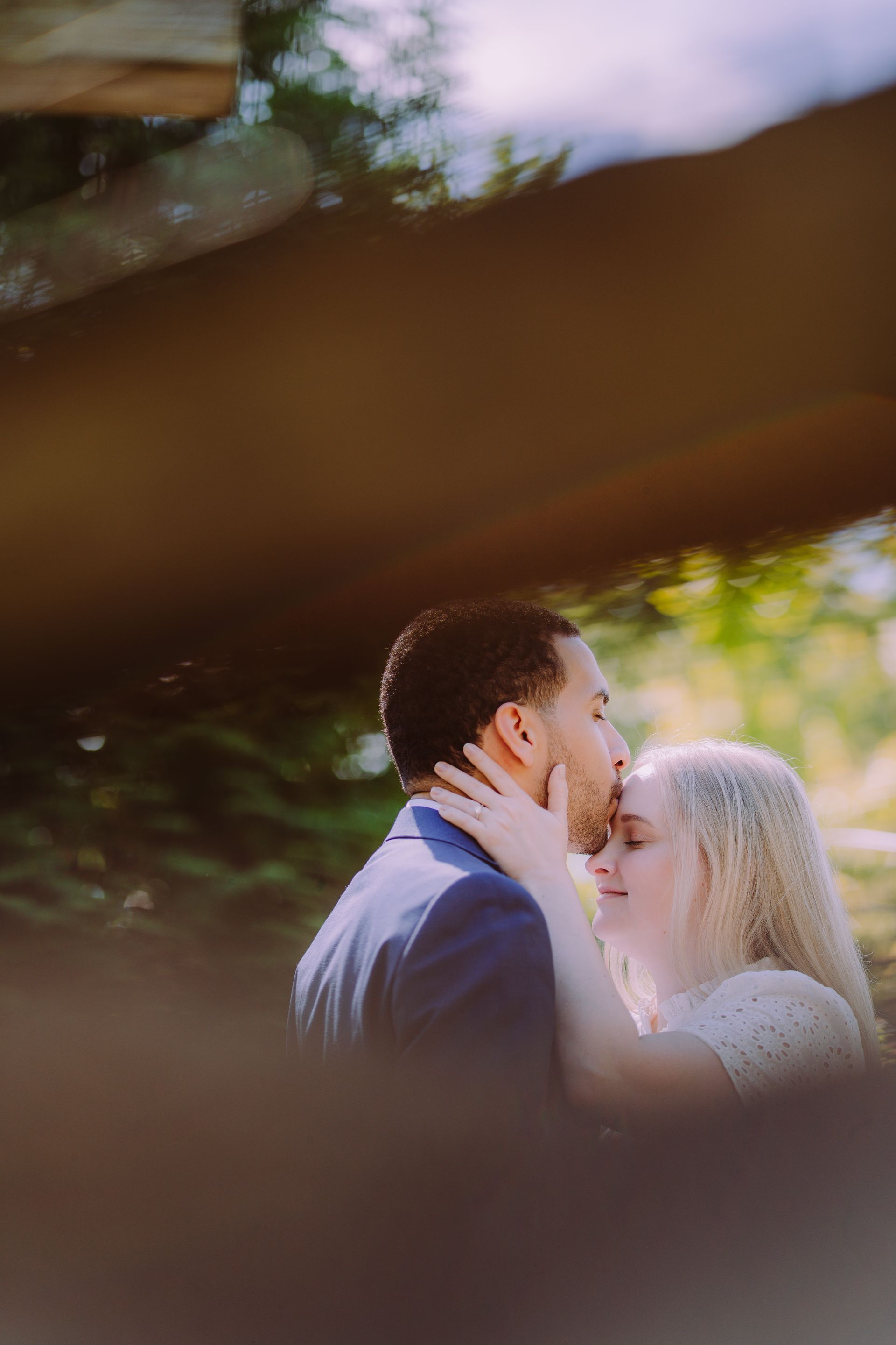 Two people share a tender forehead kiss outdoors, framed by blurred leaves and soft sunlight.