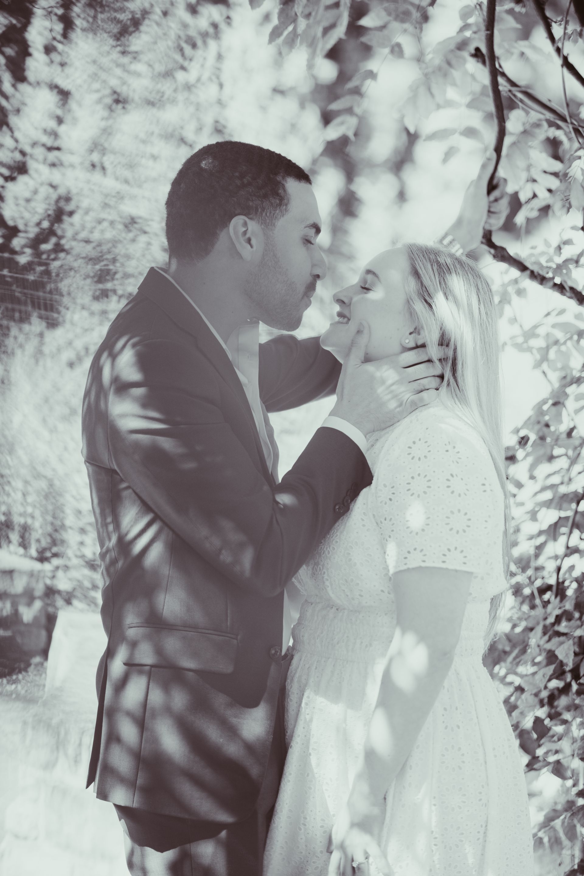 Couple in formal attire sharing a kiss outdoors by leafy branches and dappled sunlight
