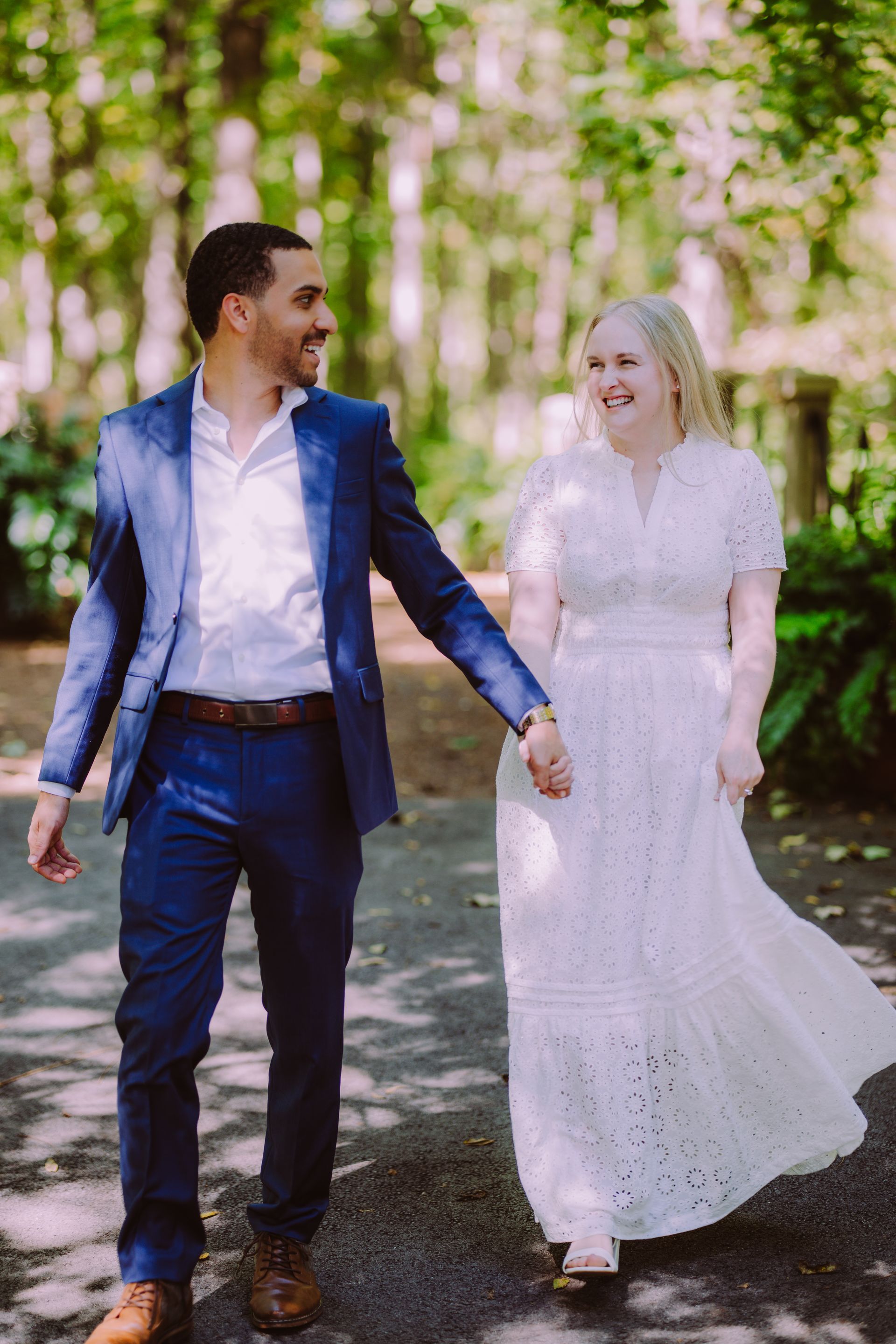 Couple holding hands and smiling on a tree-lined path, with the woman in a white dress and the man in a navy suit