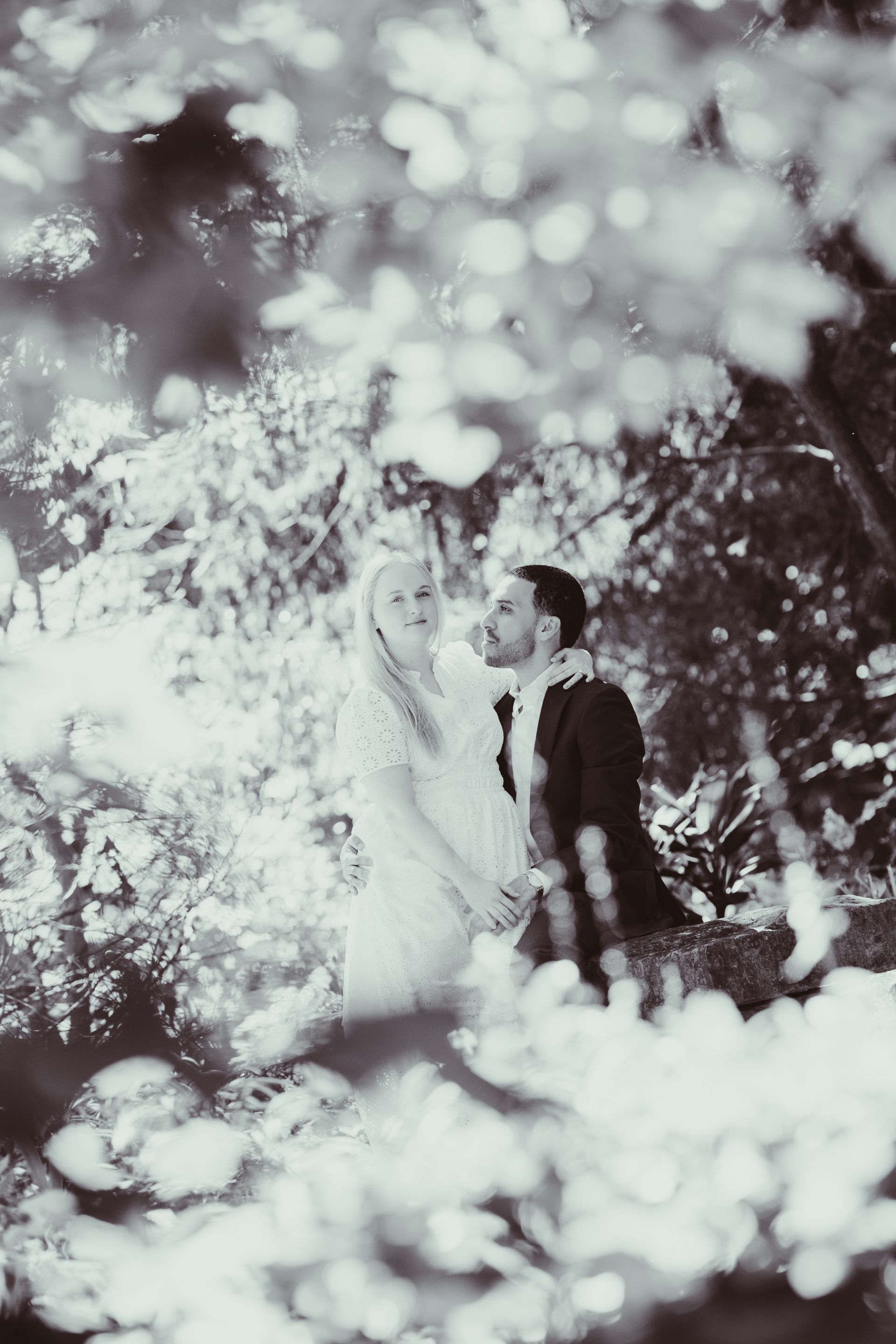 Couple standing beneath leafy branches, gazing upward in a soft, black-and-white scene