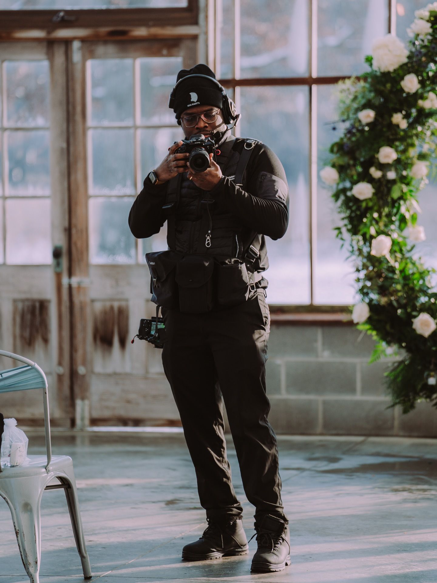 Photographer in black clothing and beanie, holding camera, indoors. Photo by Kodjo Somana