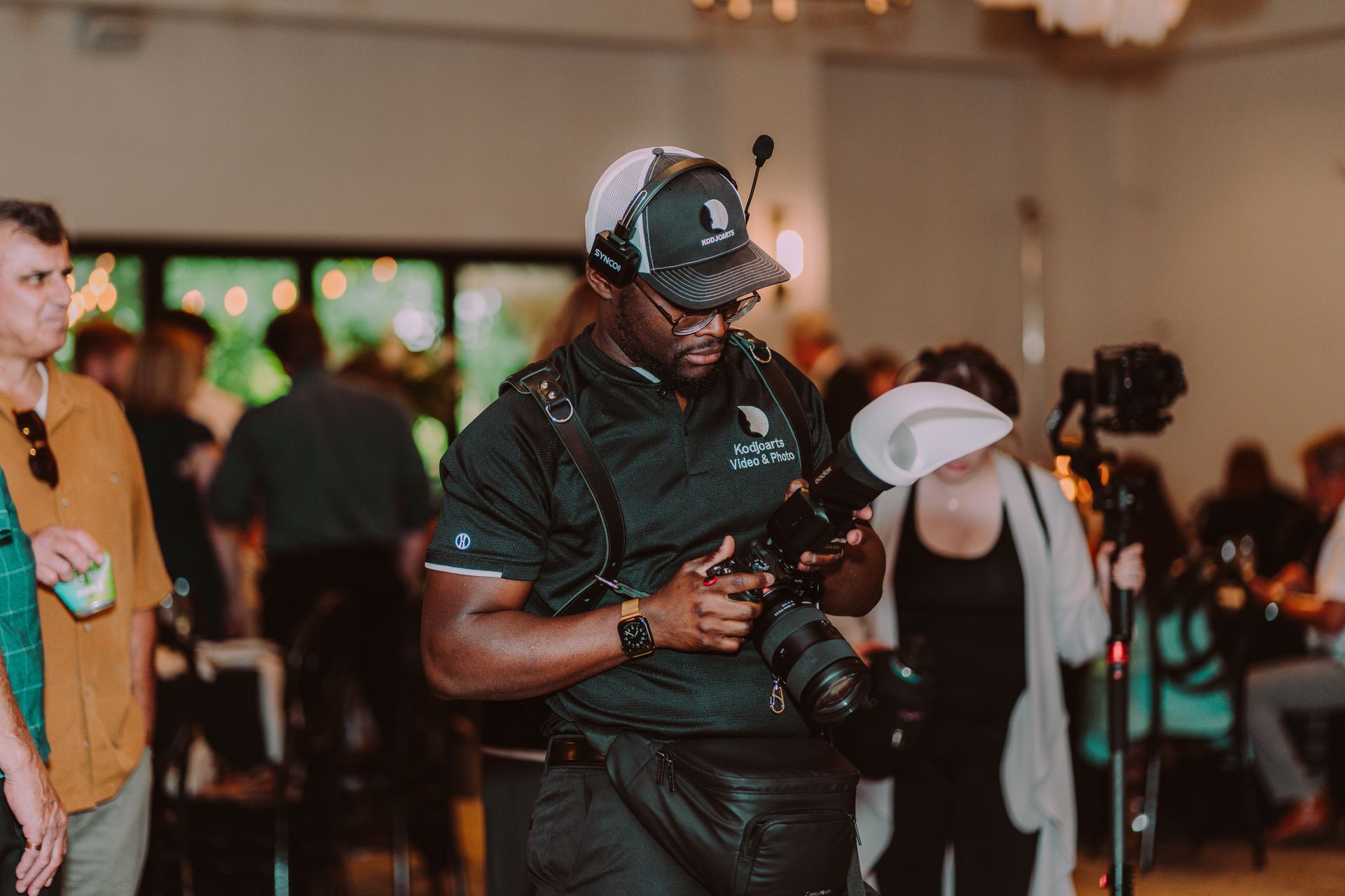 Photographer adjusting camera, wearing hat and headset, in a venue with people.