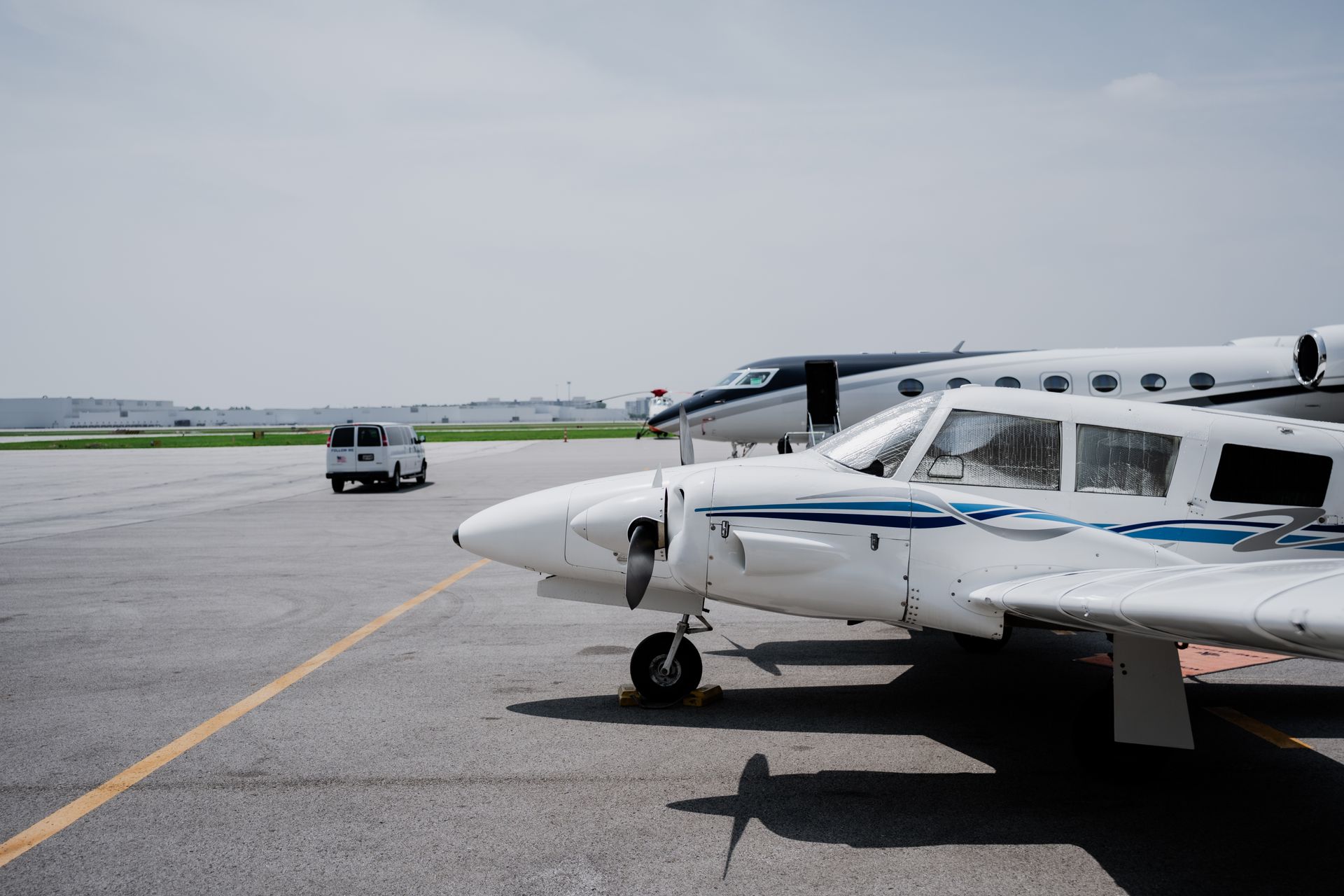 White and blue private plane parked on tarmac, other planes and a van in background.