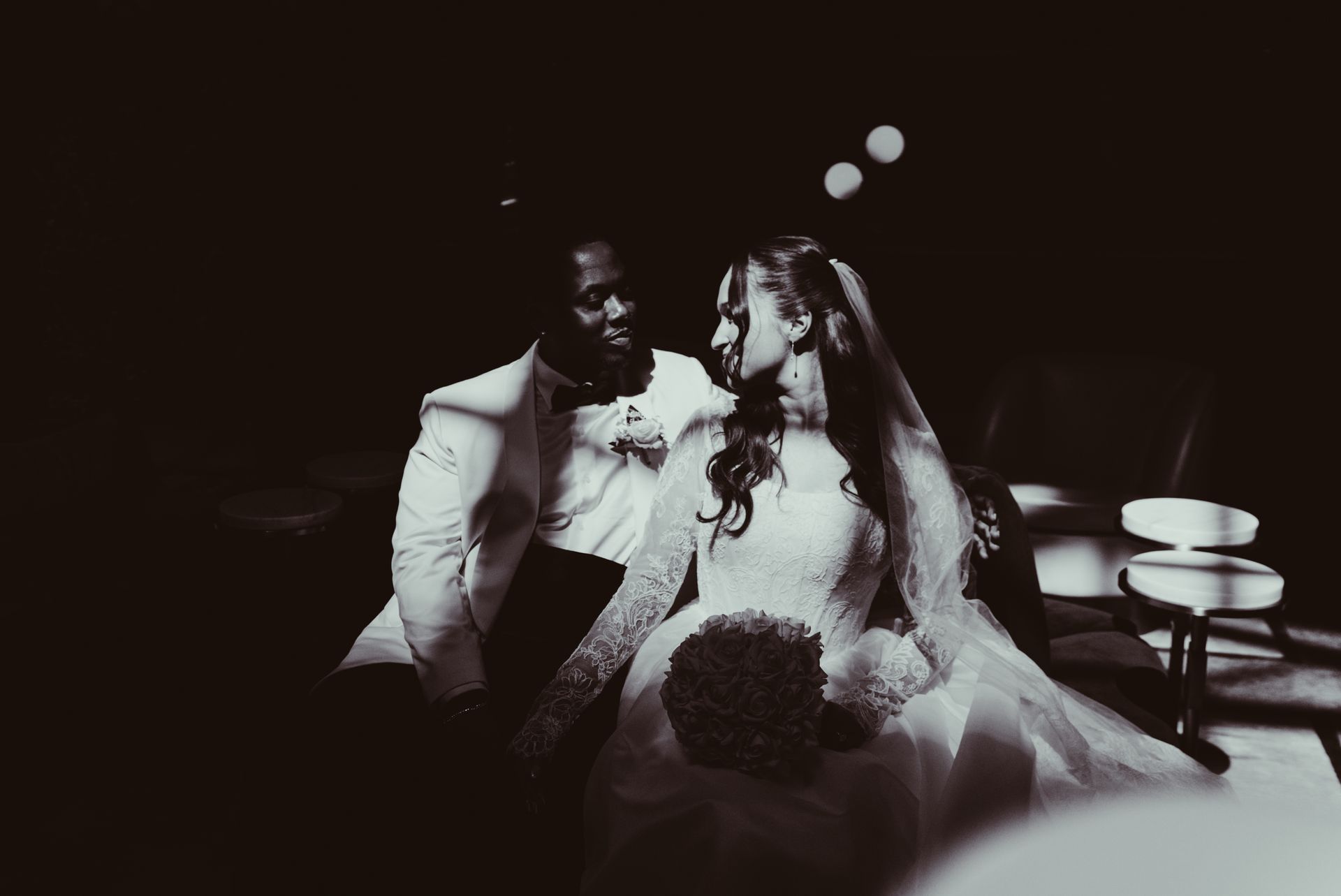 Bride and groom seated together at a dimly lit wedding reception table, smiling at each other