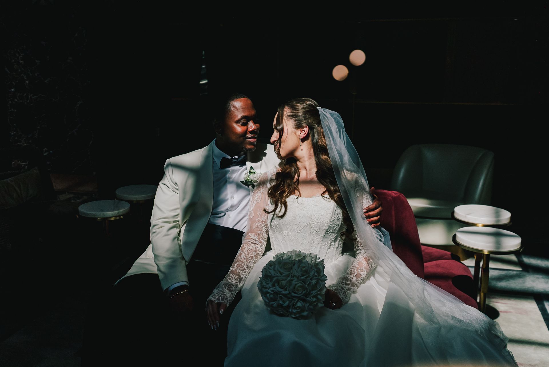 Wedding couple kissing on a couch, bride in white gown holding a bouquet in a dimly lit lounge
