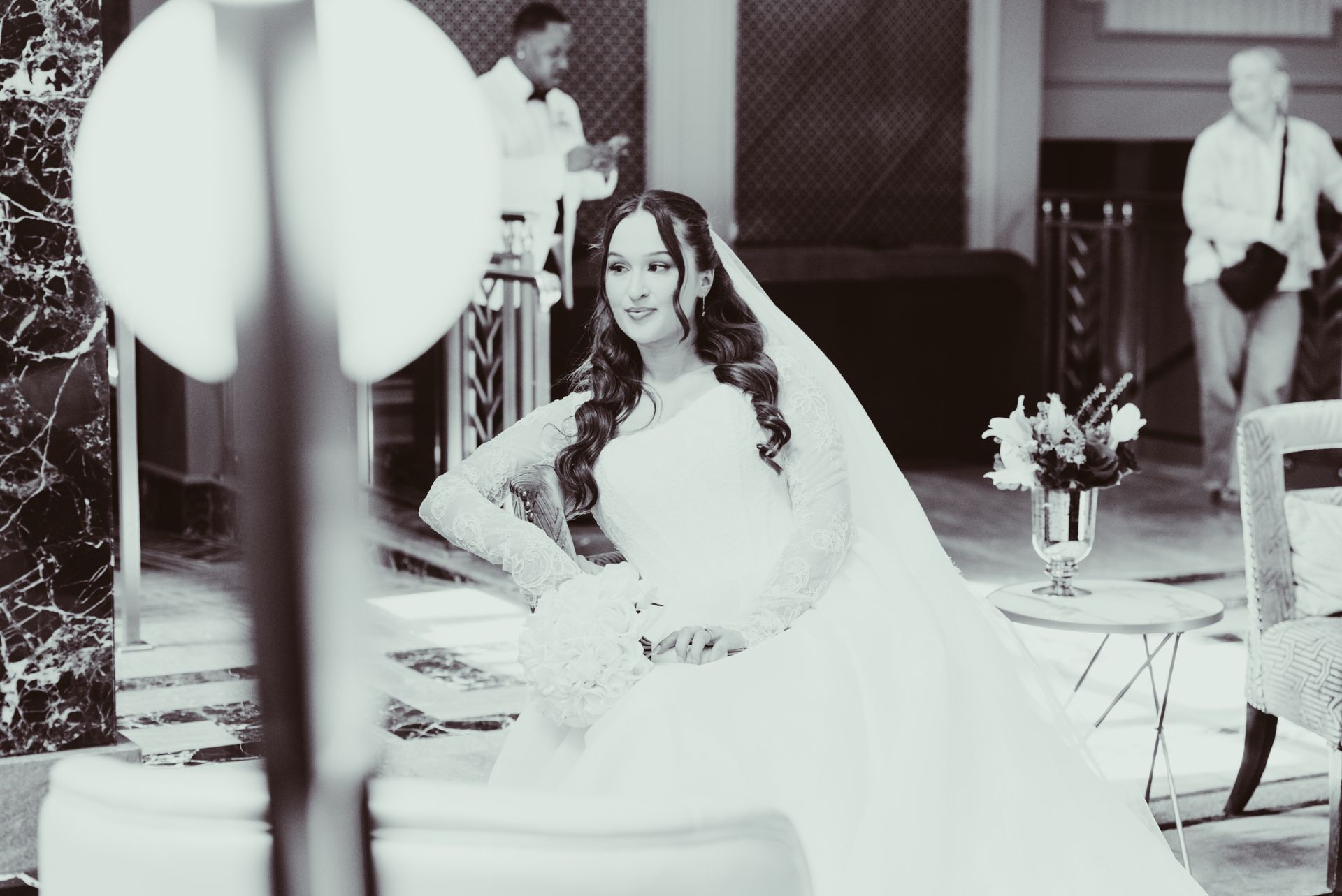 Bride in white gown seated at an indoor wedding reception, with guests and floral decor in the background
