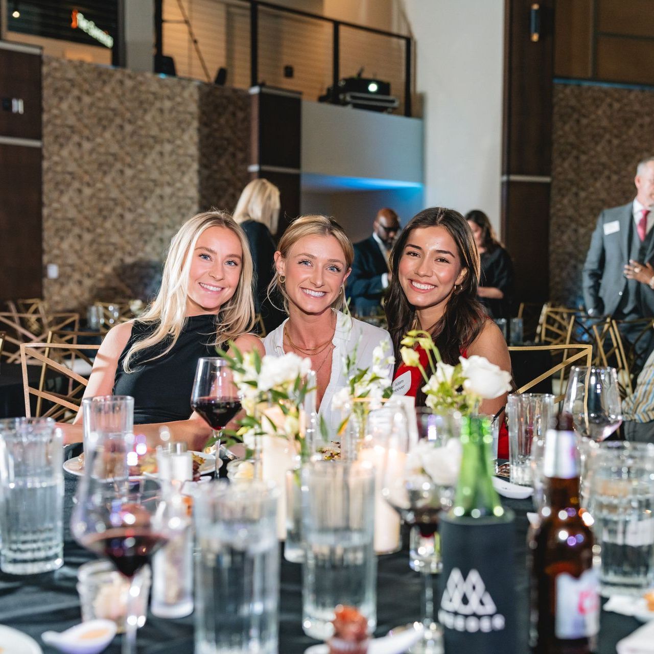 Three women are sitting at a table with wine glasses and bottles.